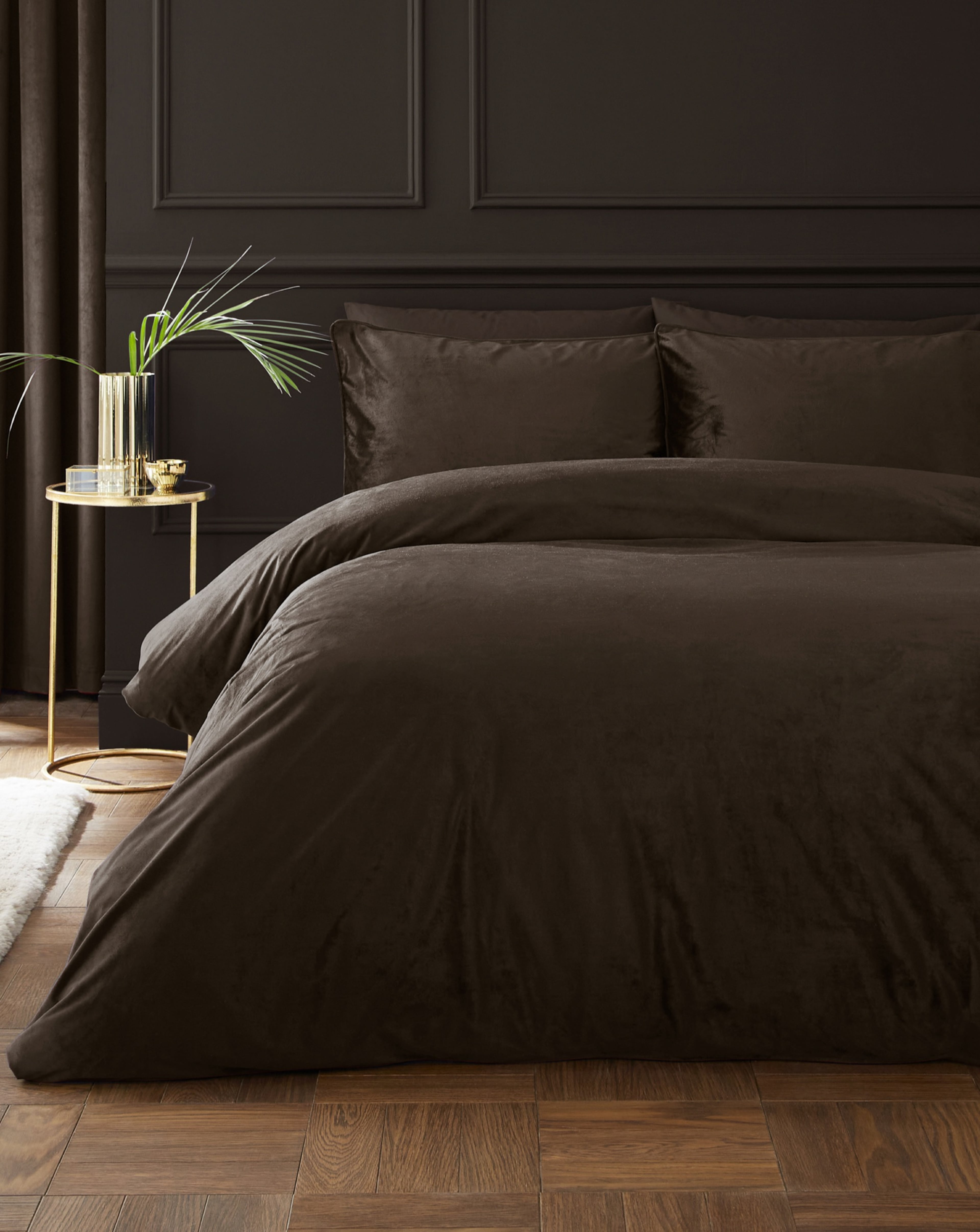 Dark, luxurious bedroom featuring a bed dressed in chocolate-coloured Catherine Lansfield bedding against a black panelled wall, with a gold side table holding a vase of green foliage and a wooden parquet floor.