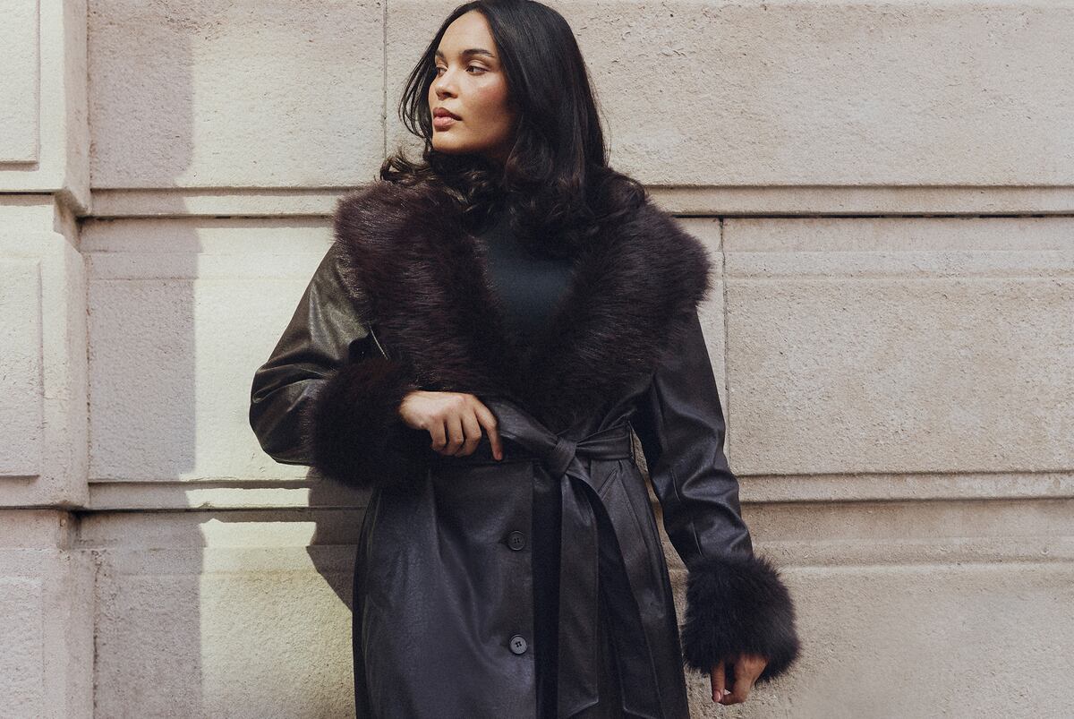 A woman with long dark hair stands against a light-coloured stone wall, wearing a dark coat with a plush faux-fur collar and cuffs.