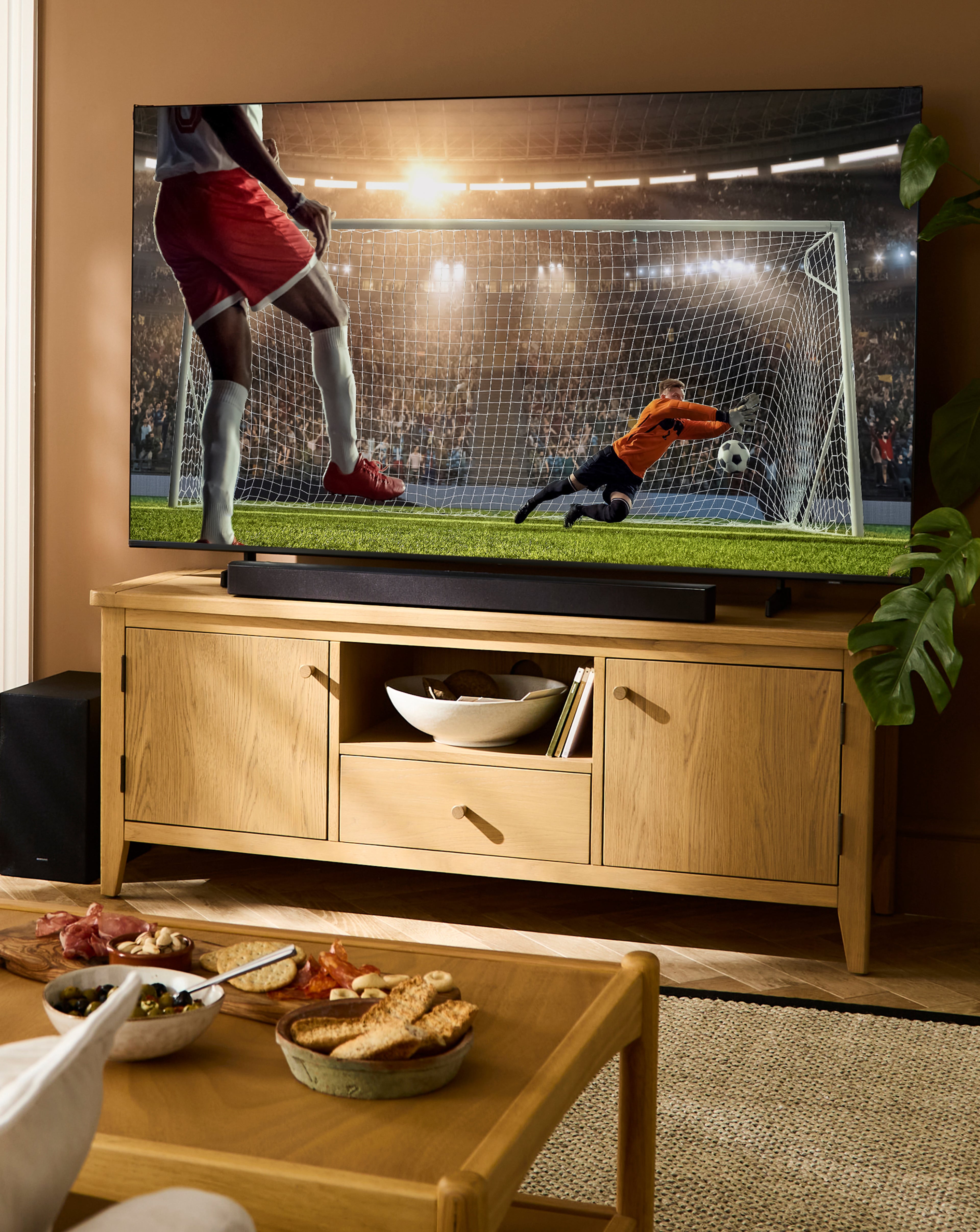 Living room scene with a large TV on a wooden TV unit, showing a goalkeeper diving for a football.