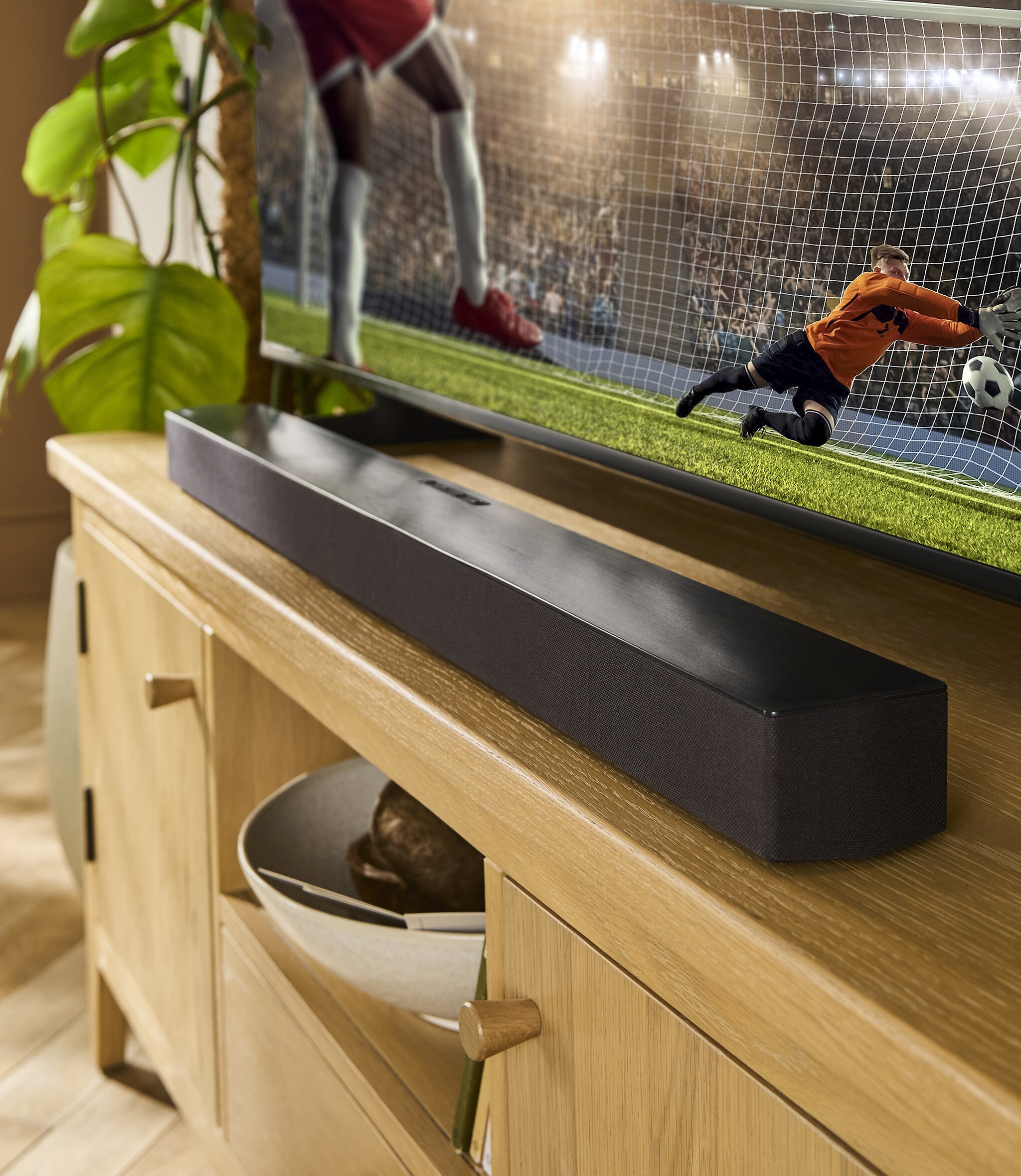 A black soundbar on a TV unit in front of a TV on displaying a football match in action.