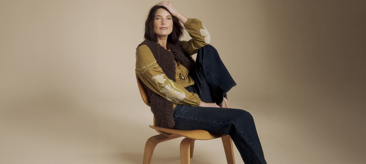 A woman sat on a wooden chair wearing a brown wooly gilet and mustard coloured top and dark blue jeans.