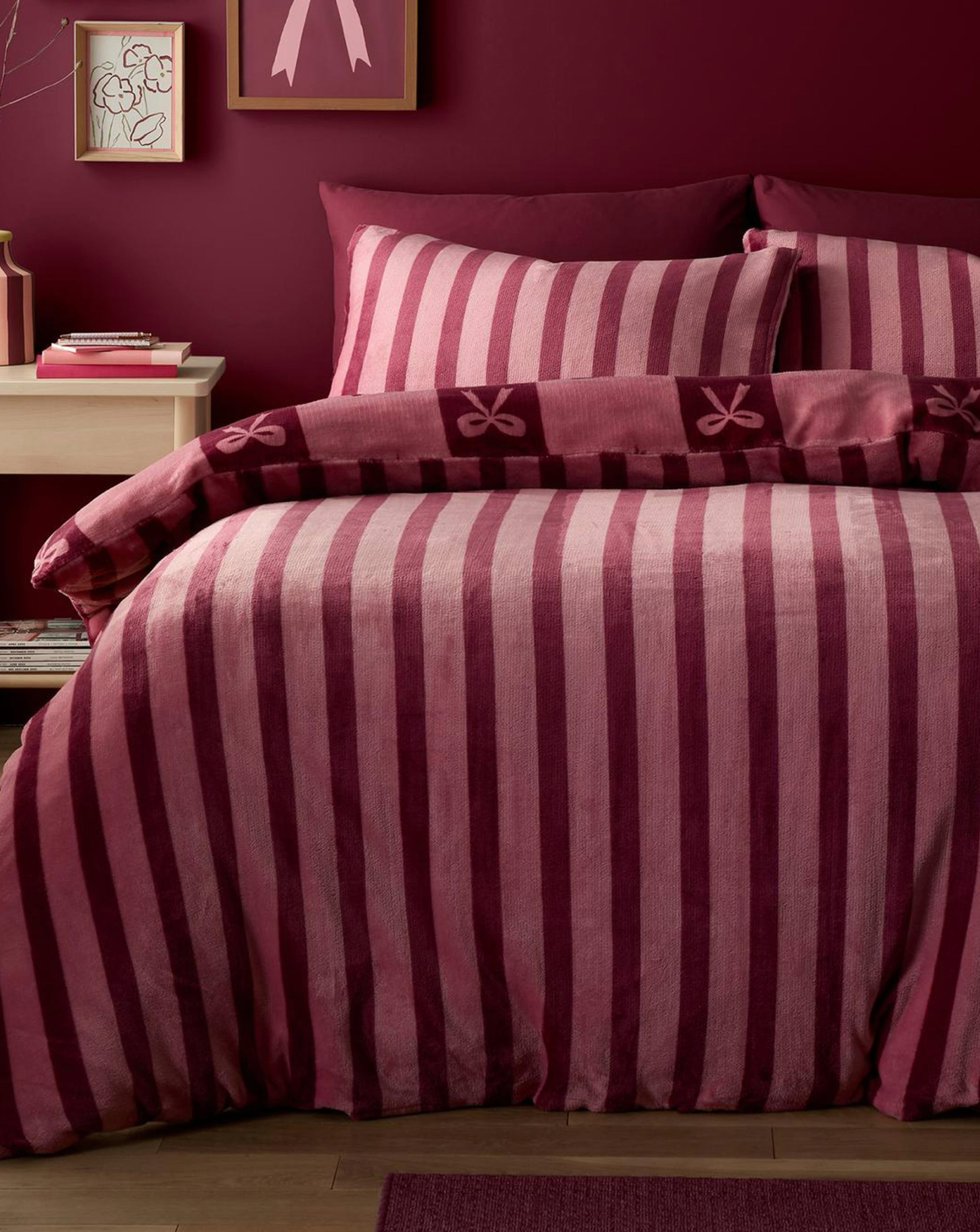 A bed dressed in pink and burgundy striped bedding with a bow motif, set against a deep red wall with framed artwork.