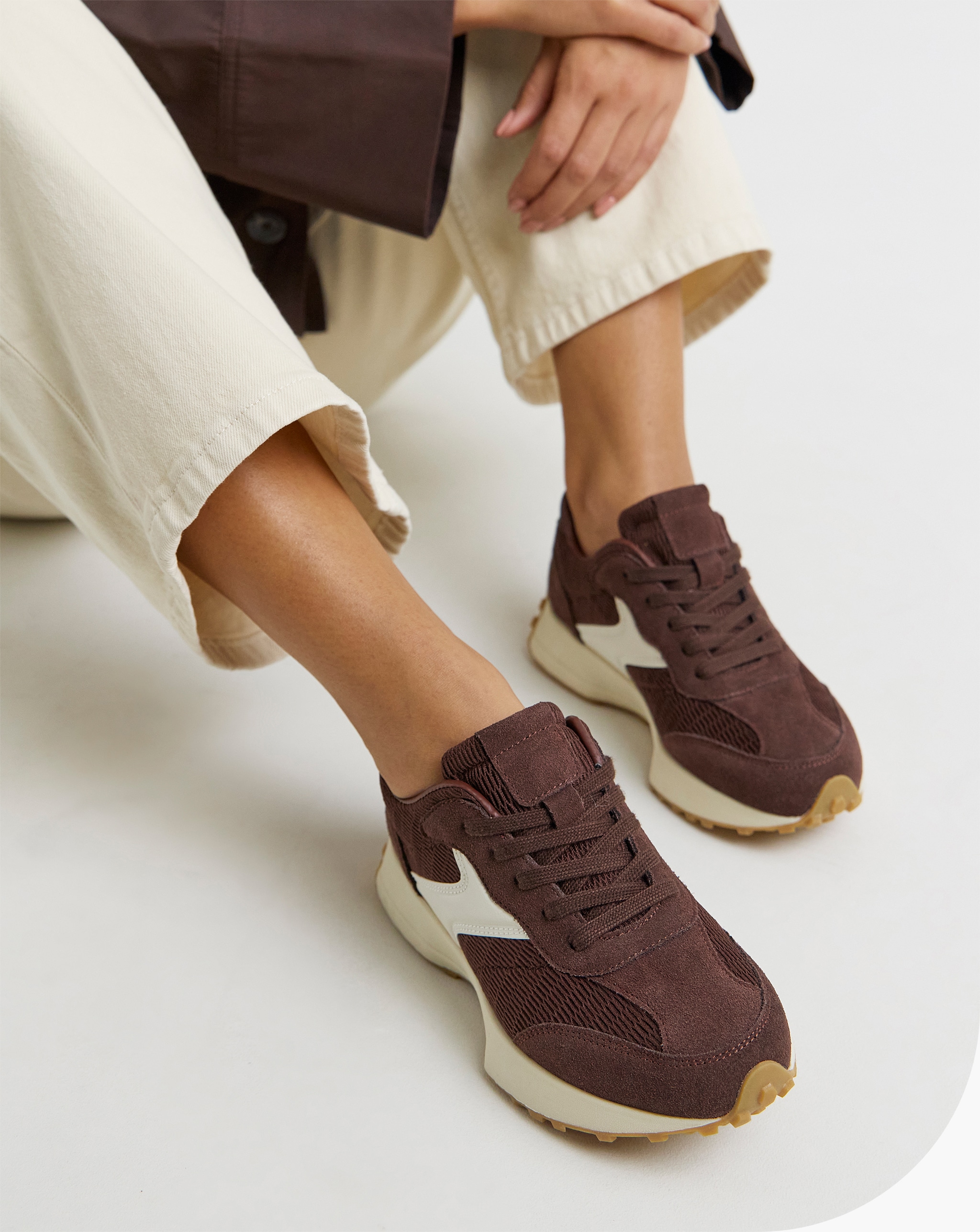 A close up of a person sitting wearing brown Akira Suede Retro Runner Trainers