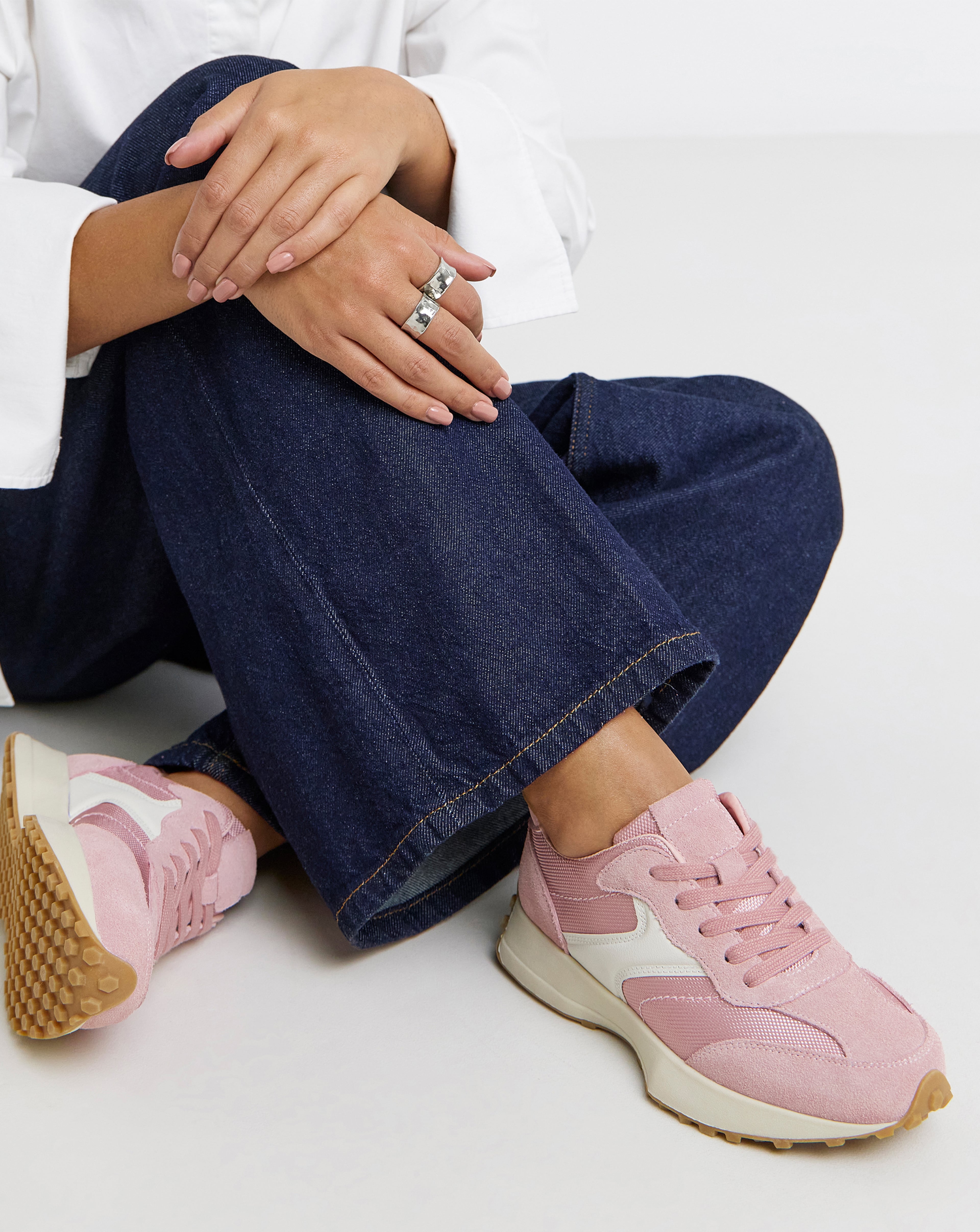 Close-up of a person sat down, wearing pink Akira Suede Retro Runner Trainers and blue jeans.