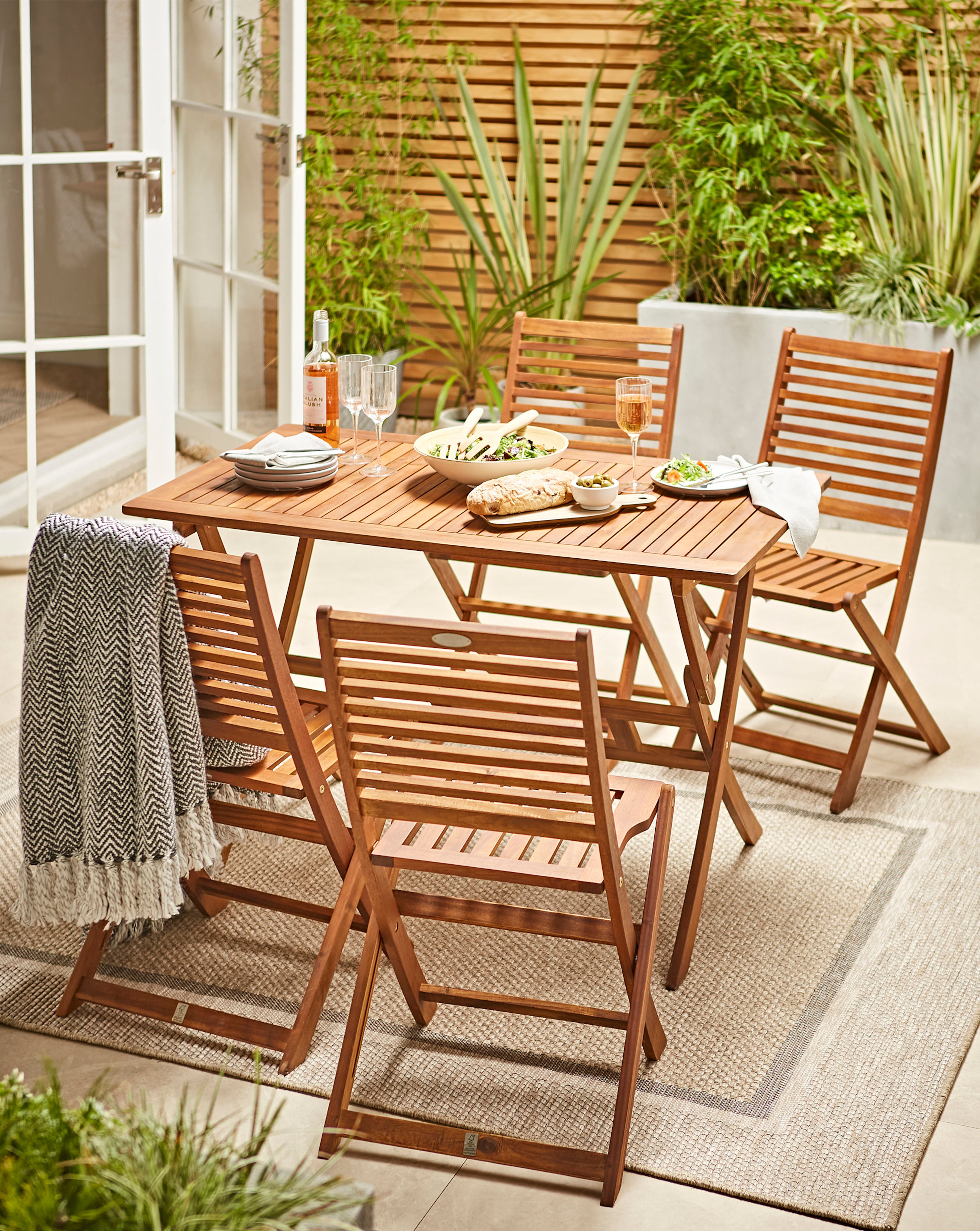 Wooden garden table and chairs set with food and wine, beside an open patio door and plants.