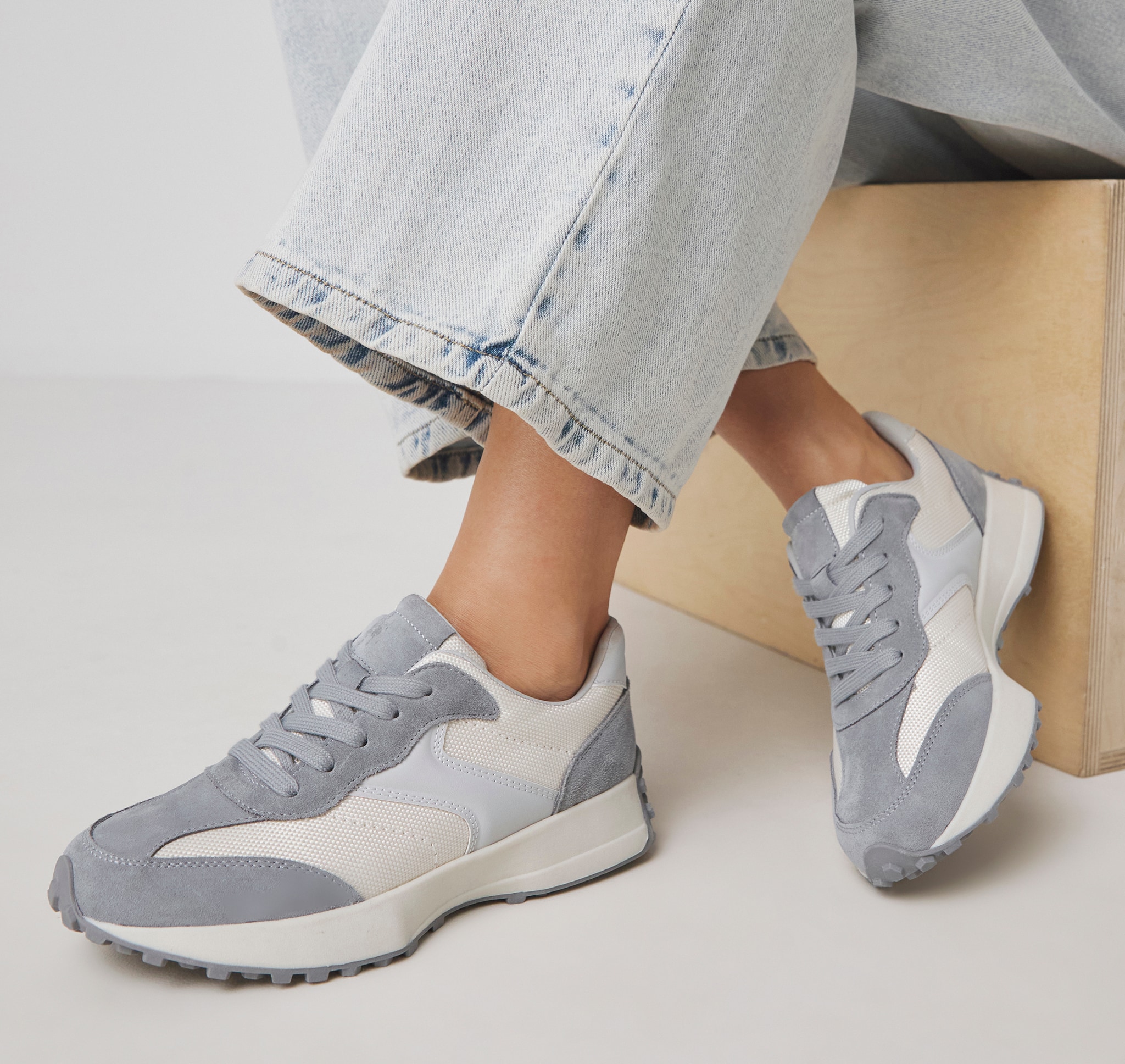 A person sat on a wooden box wearing pale blue jeans, no sock and grey panelled trainers
