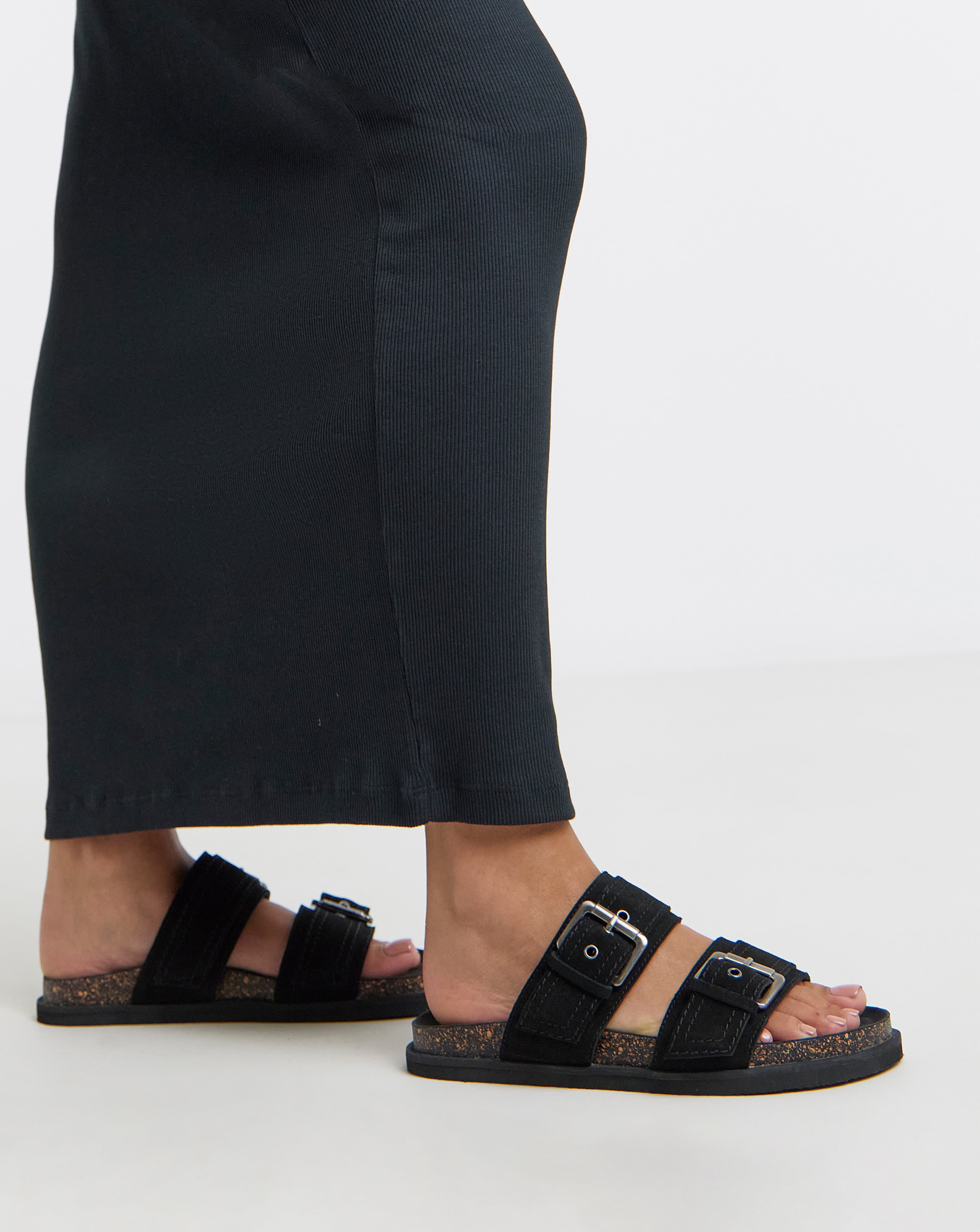Lower-leg view of black ribbed trousers paired with black buckle sandals against a light background.