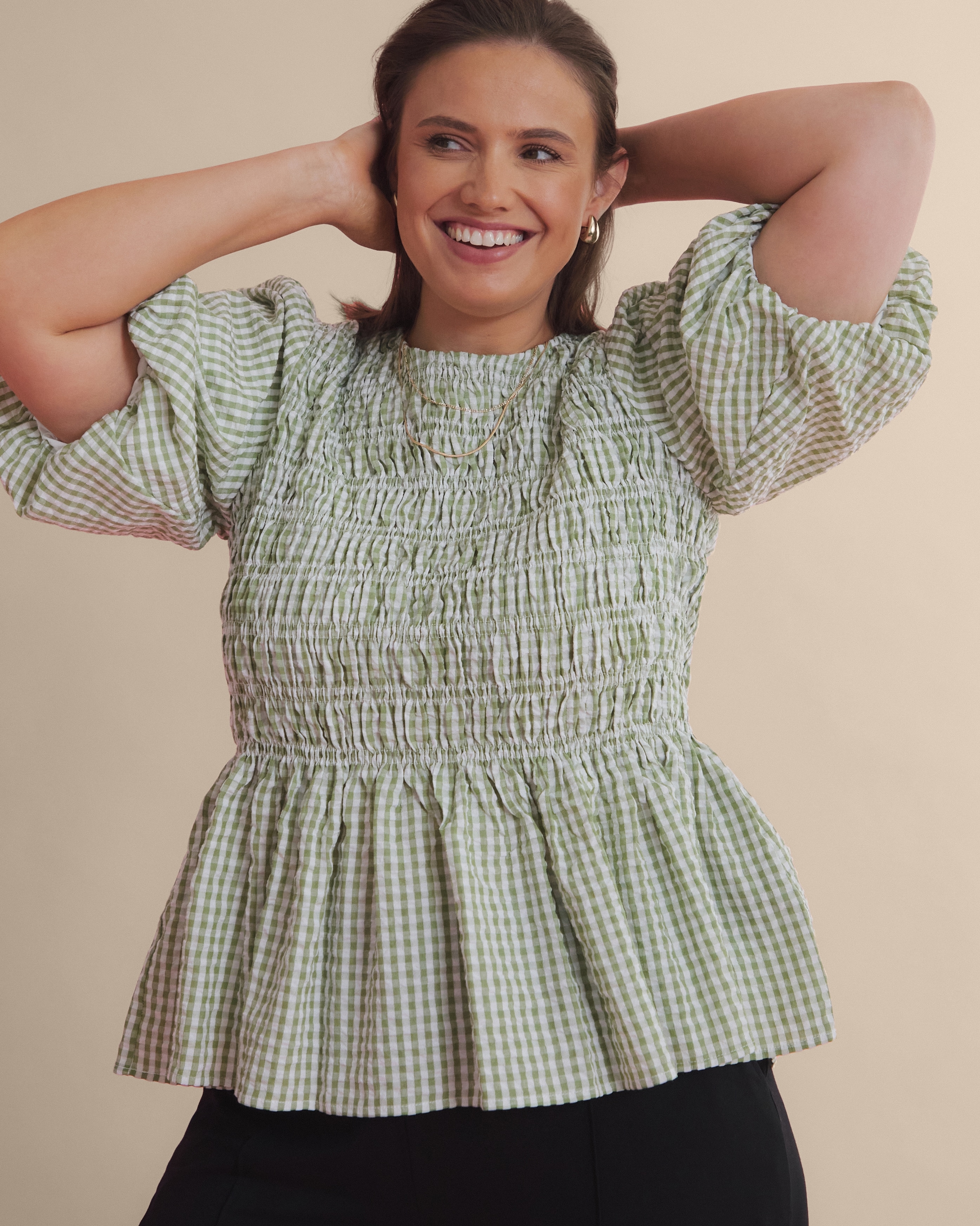 Confident woman wearing a stylish green gingham blouse smiling warmly in a studio setting. Simple and visually appealing image emphasizing fashion, comfort, and joyful expression, perfect for retail or lifestyle themes.