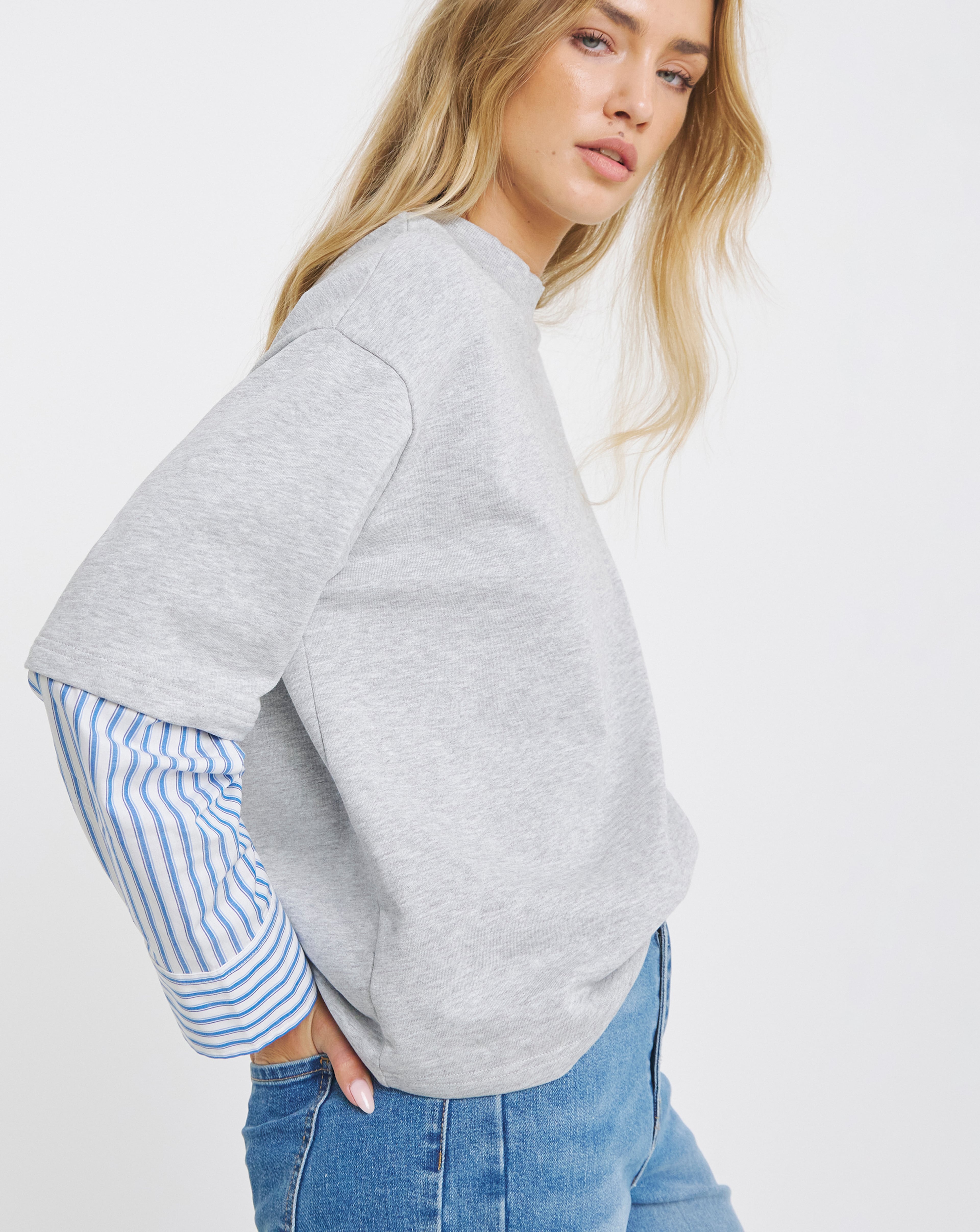Person wearing a light grey short-sleeve sweatshirt layered over a blue-and-white striped shirt, paired with blue jeans.