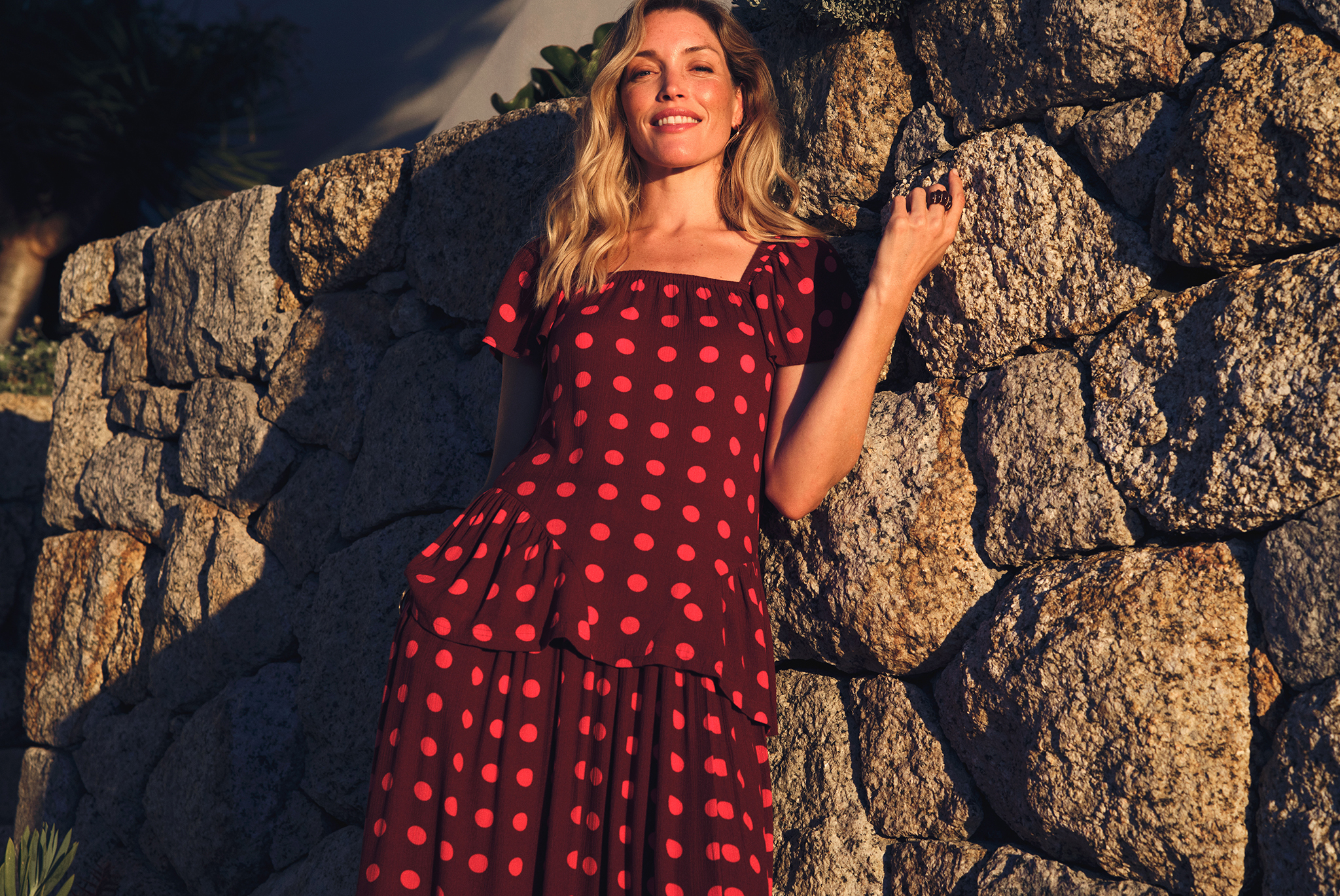 Person in a red, polka‑dot dress standing against a sunlit stone wall outdoors, with warm light casting shadows.
