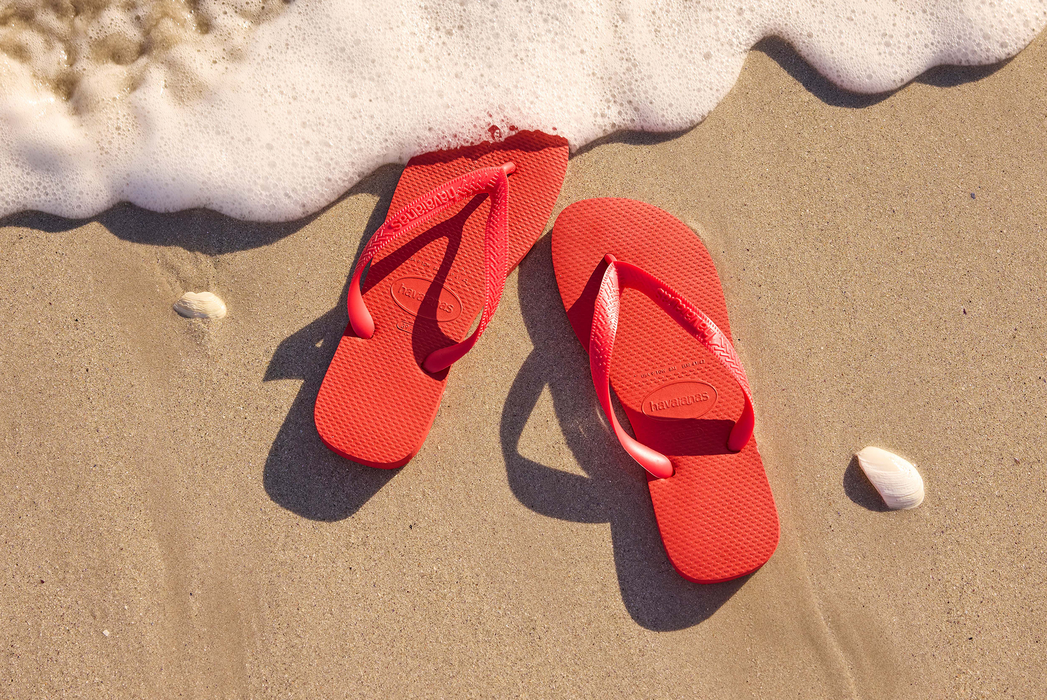 Bright red flip-flops on sandy beach near foamy shoreline with small seashells.