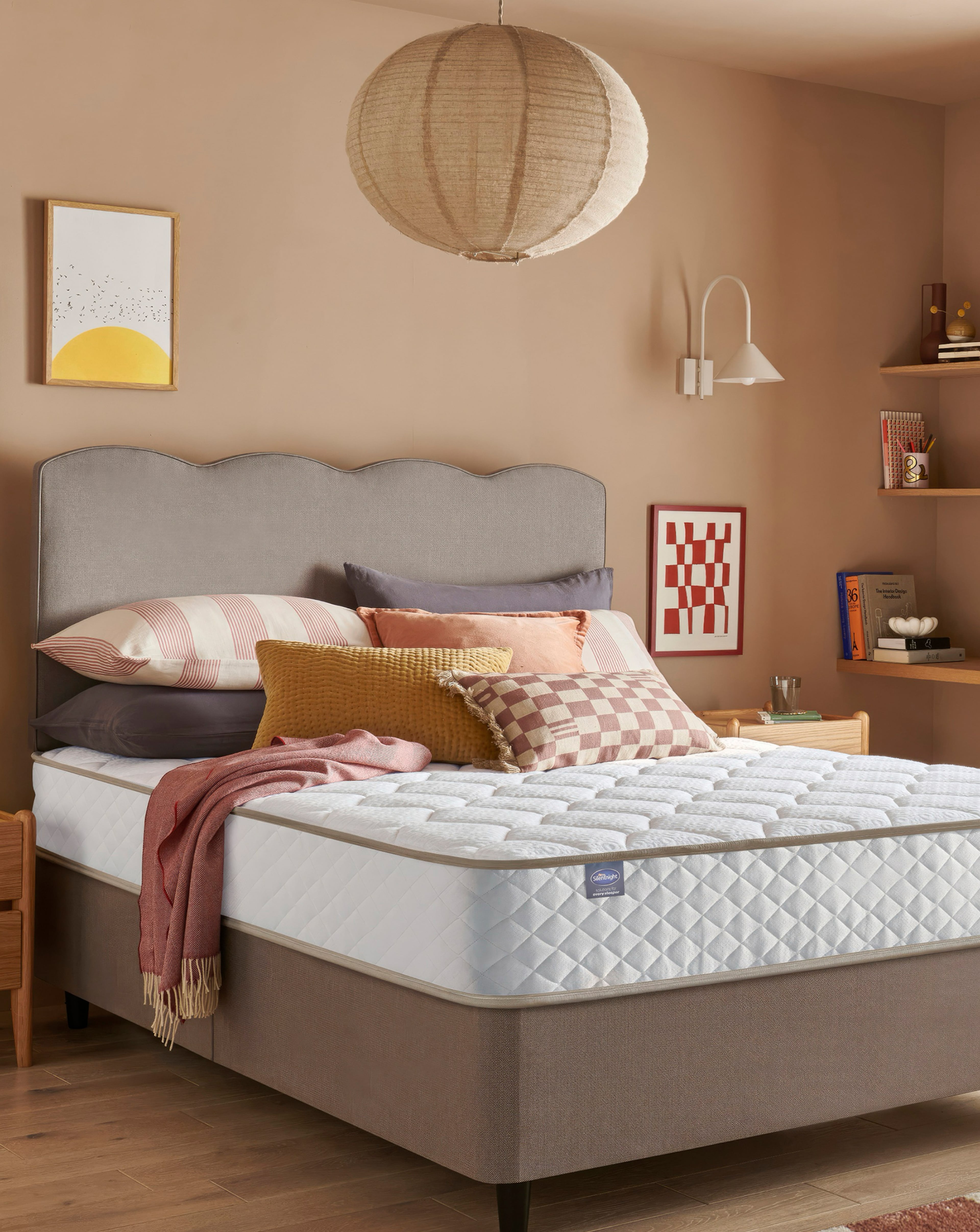 Stylish bedroom featuring a upholstered bed with a quilted Silentnight mattress, layered cushions and throws, warm-toned walls, floating shelves, and soft ambient lighting.