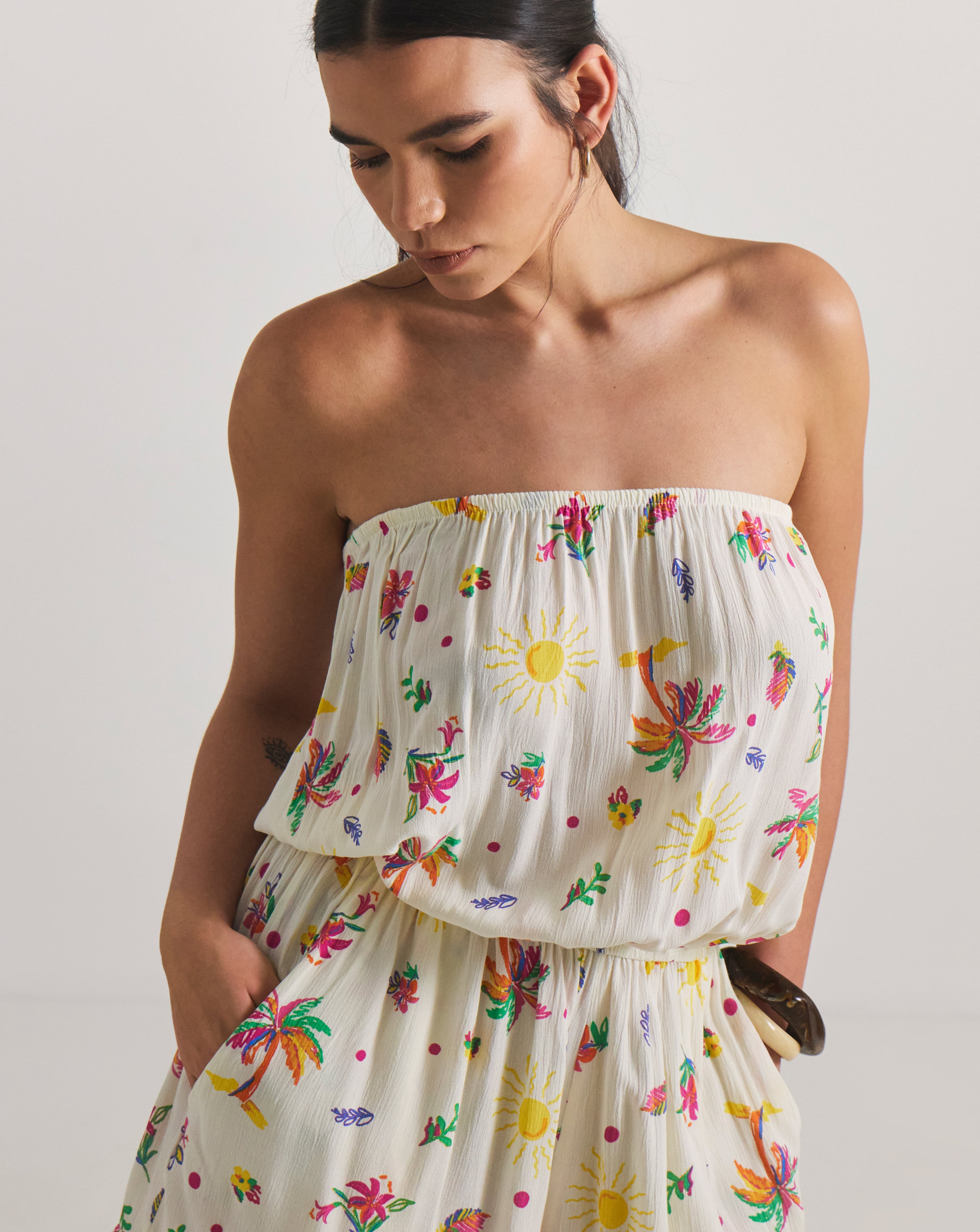 Person wearing a strapless cream dress with colourful floral and sun prints, with hands in pockets.