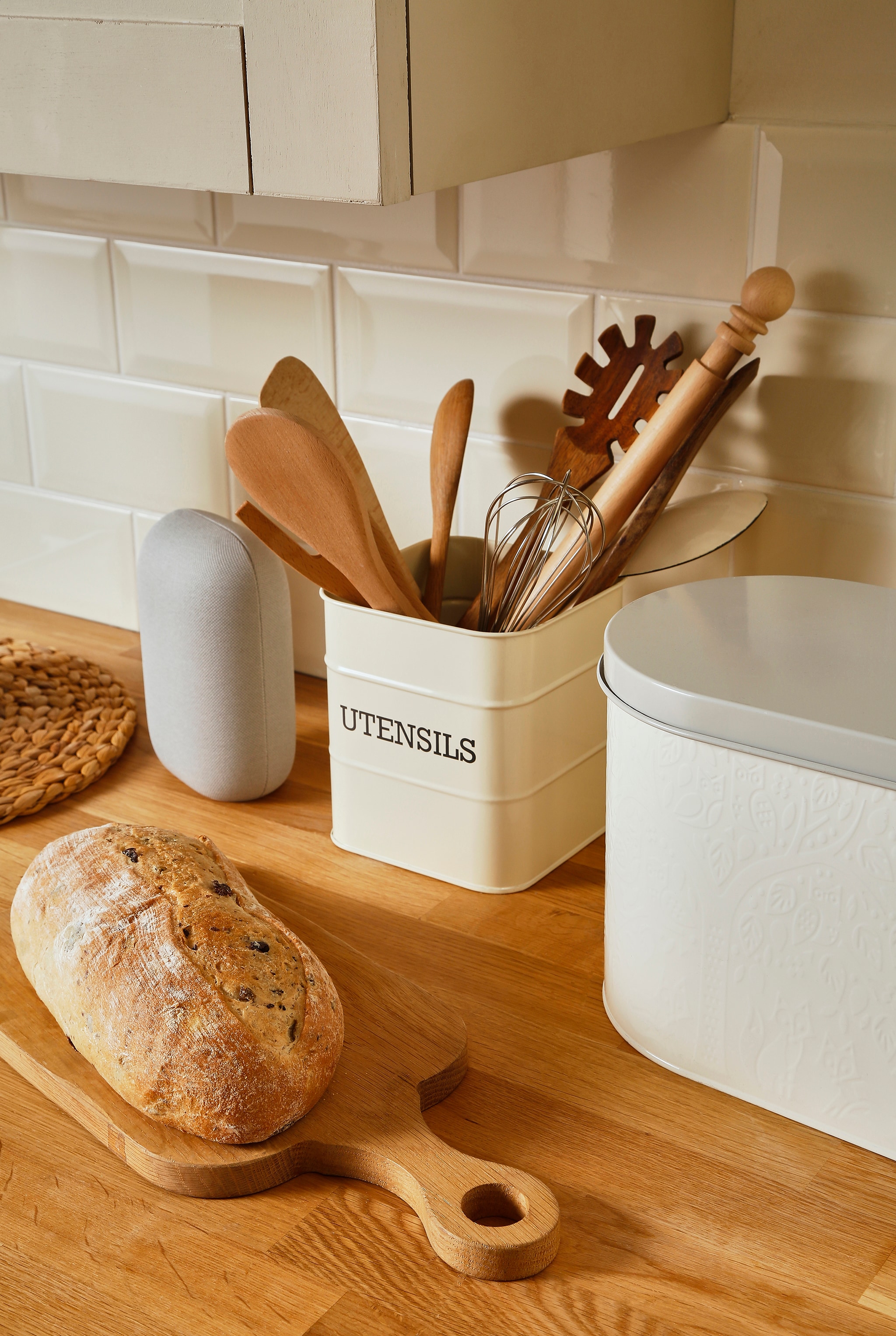 A kitchen worktop with some fresh bread on a chopping board. Utensils in a metal rectangular container displaying the word utensils on it written in captials.