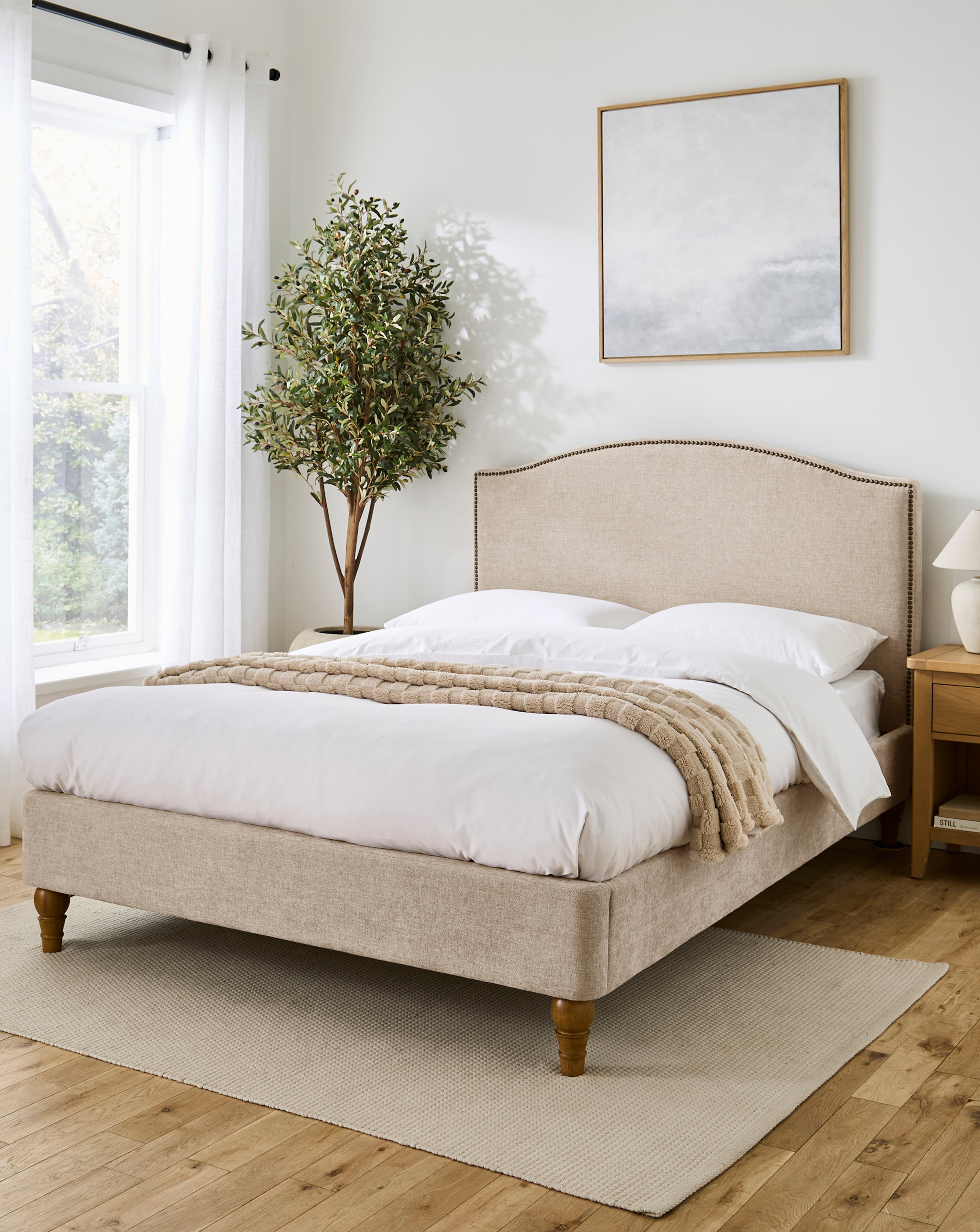 Bright, neutral-toned bedroom featuring an upholstered bed with white bedding, a throw blanket, bedside table, framed wall art, and a potted tree by a large window.
