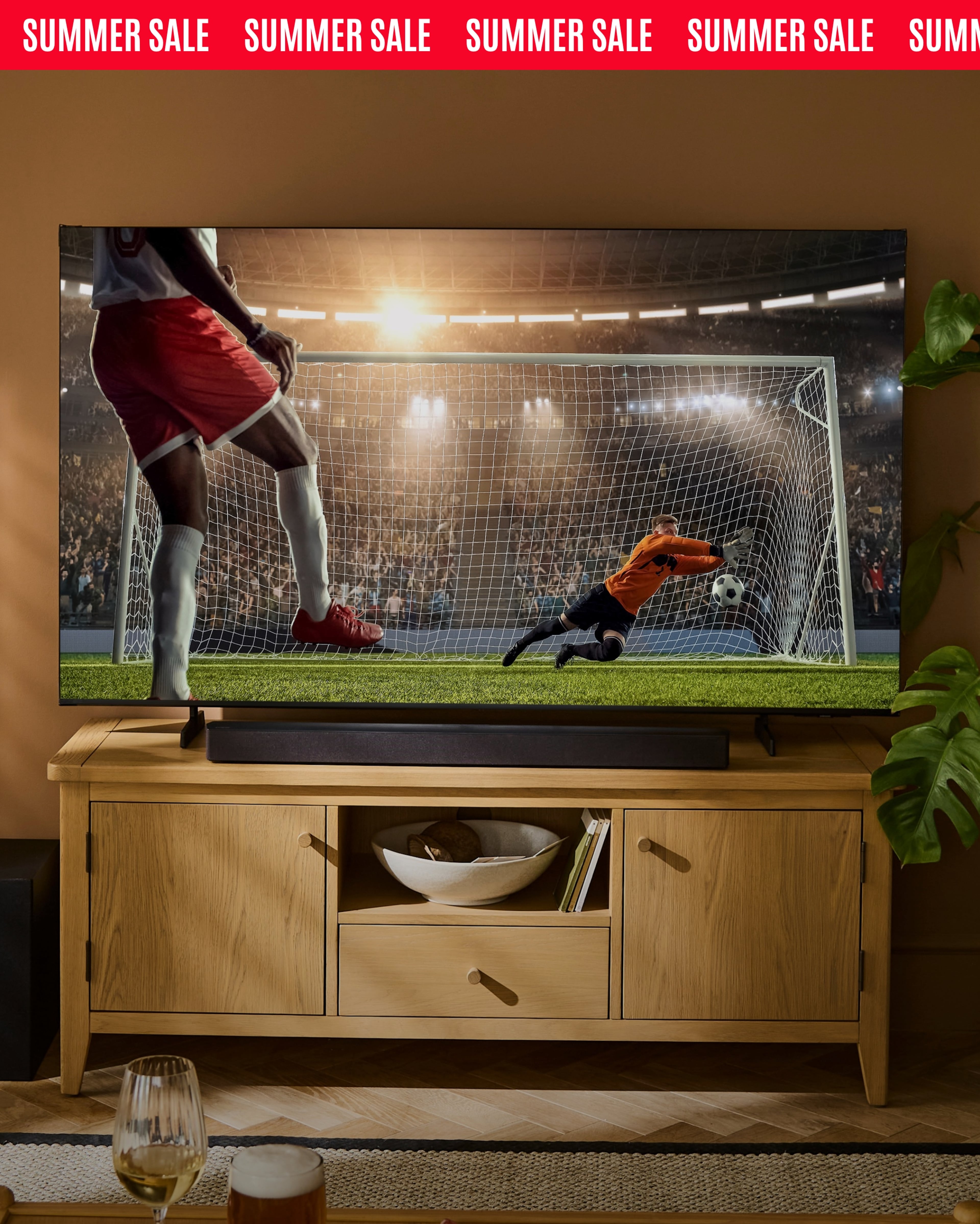Summer sale banner showing a large TV displaying a football penalty kick in a living room setting.