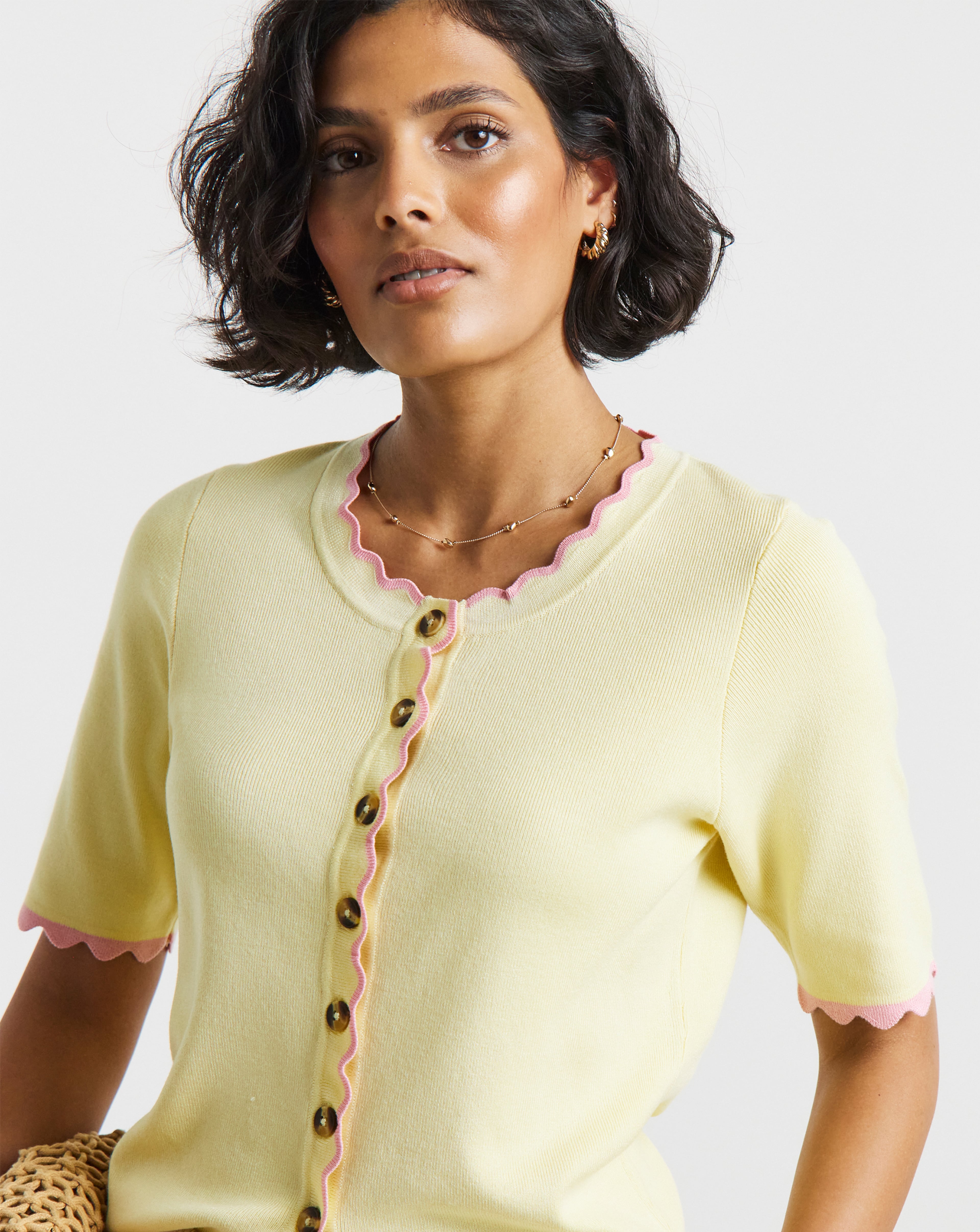 Close-up of a person wearing a pale yellow short-sleeve knitted cardigan with pink scalloped trim, tortoiseshell-style buttons, and gold jewellery. 