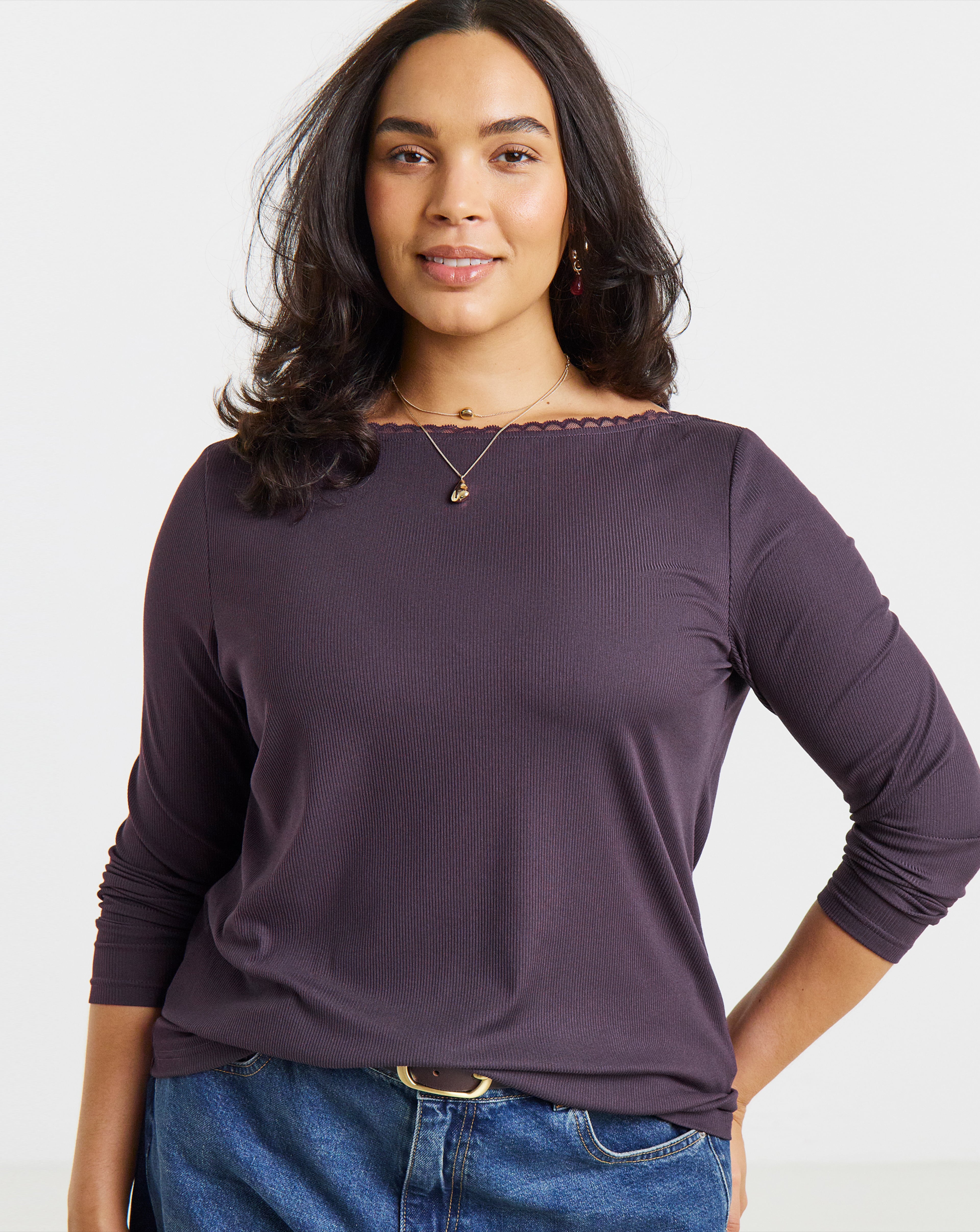 Studio portrait of a person wearing a long-sleeved ribbed top, layered necklaces, and jeans against a light grey background.