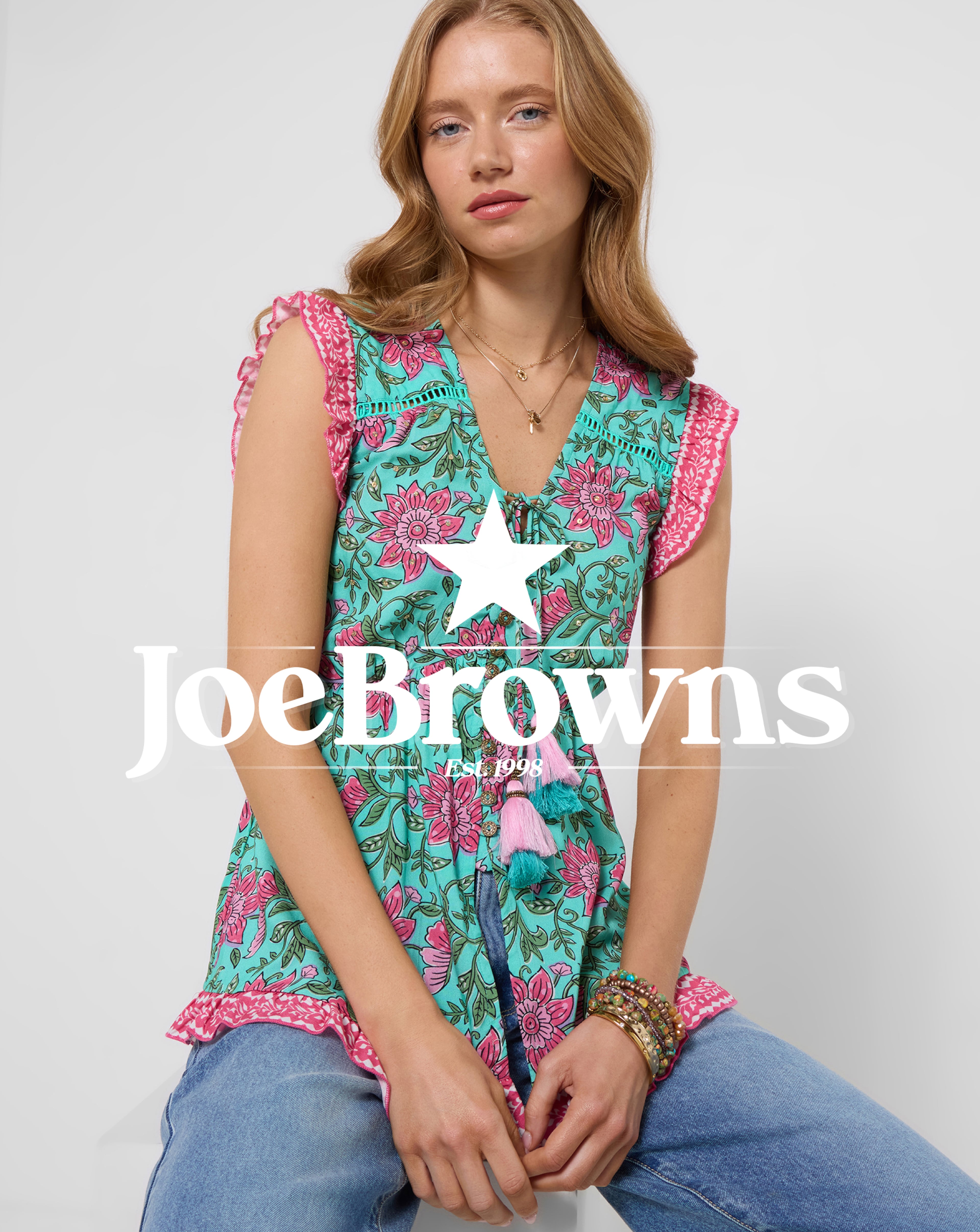 Person seated in jeans and a Joe Browns turquoise floral sleeveless blouse with pink ruffle details and tassel ties.
