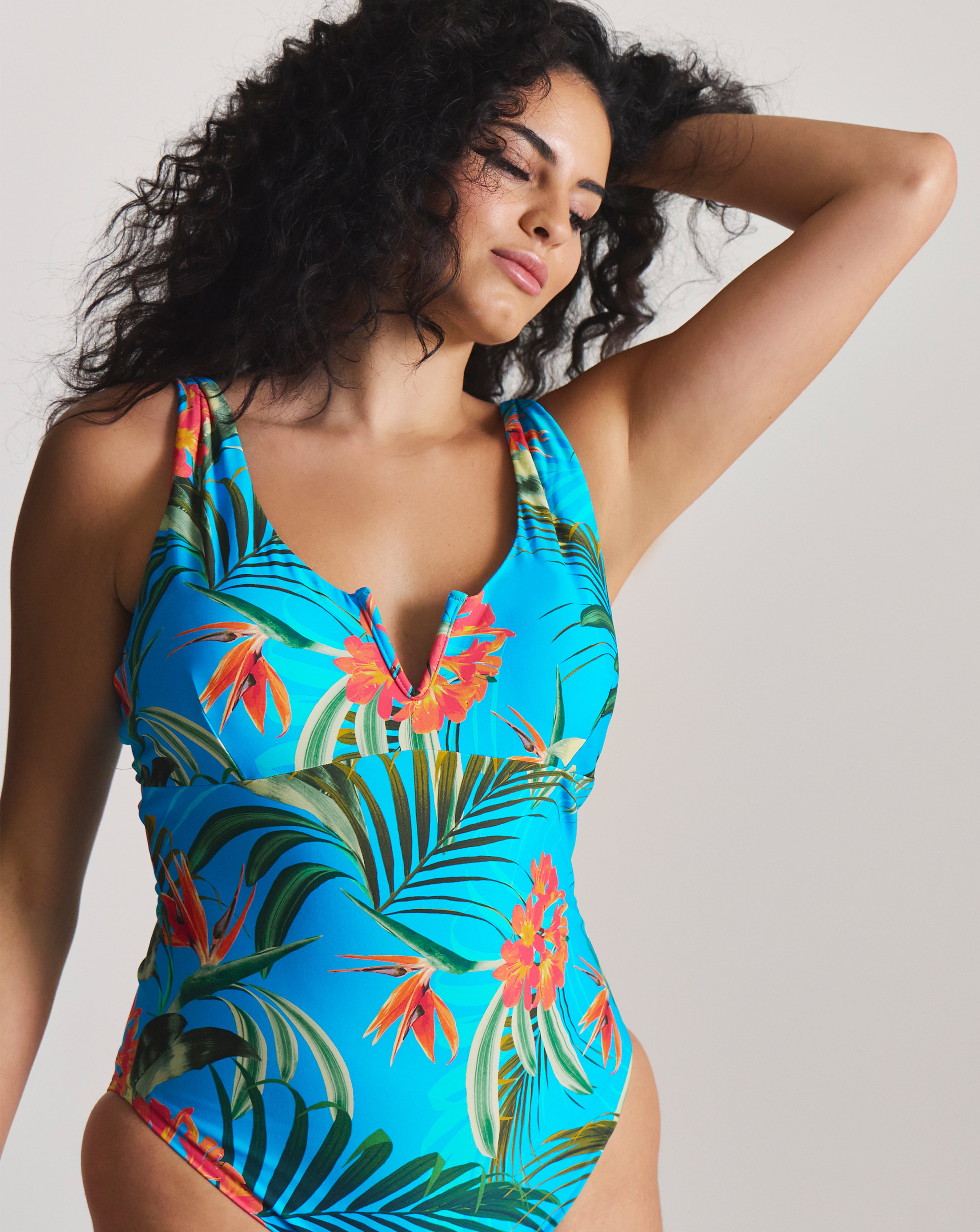 Person modelling a bright blue tropical-print swimsuit with colourful floral and palm-leaf design.