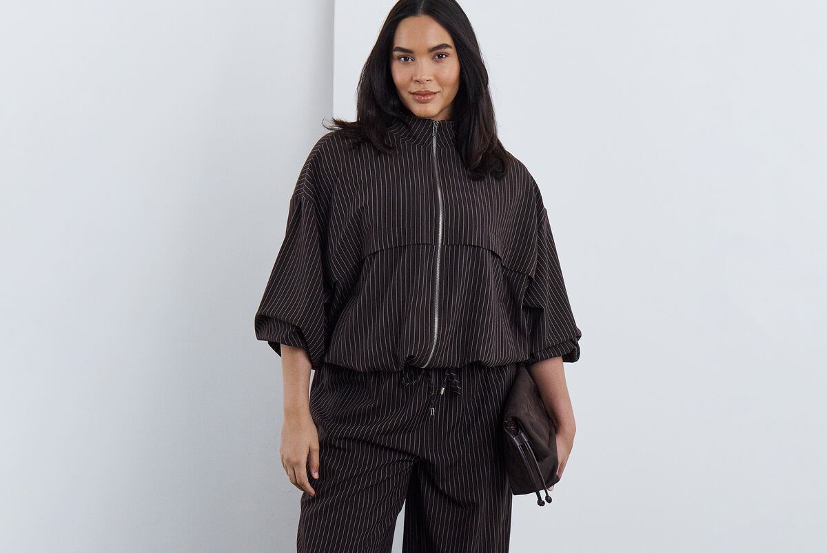 A person standing against a white wall wearing a dark brown pinstripe matching top and trousers.
