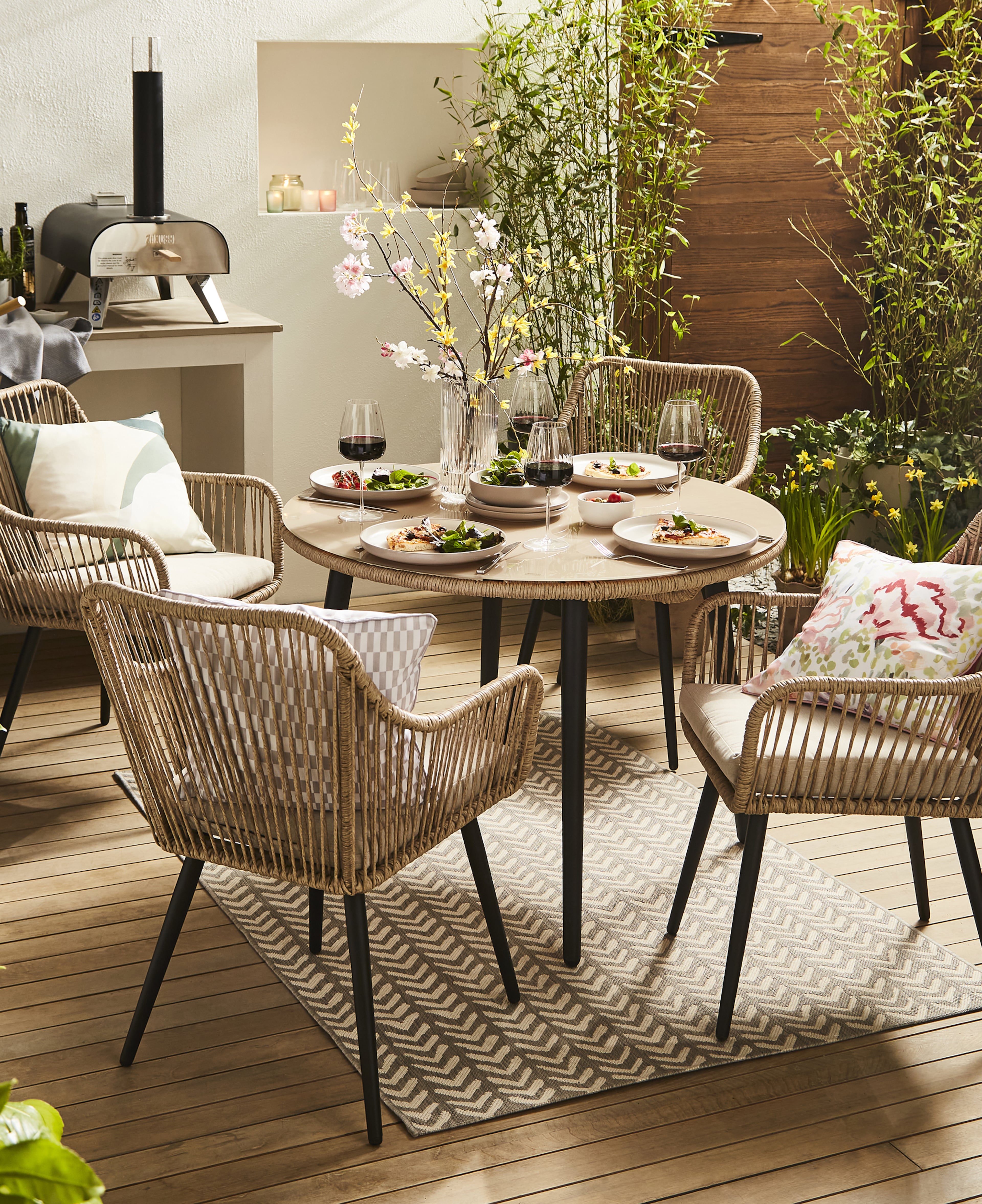 Outdoor patio dining set with a round table, woven chairs and cushions, place settings with food and wine, surrounded by greenery and a compact pizza oven.