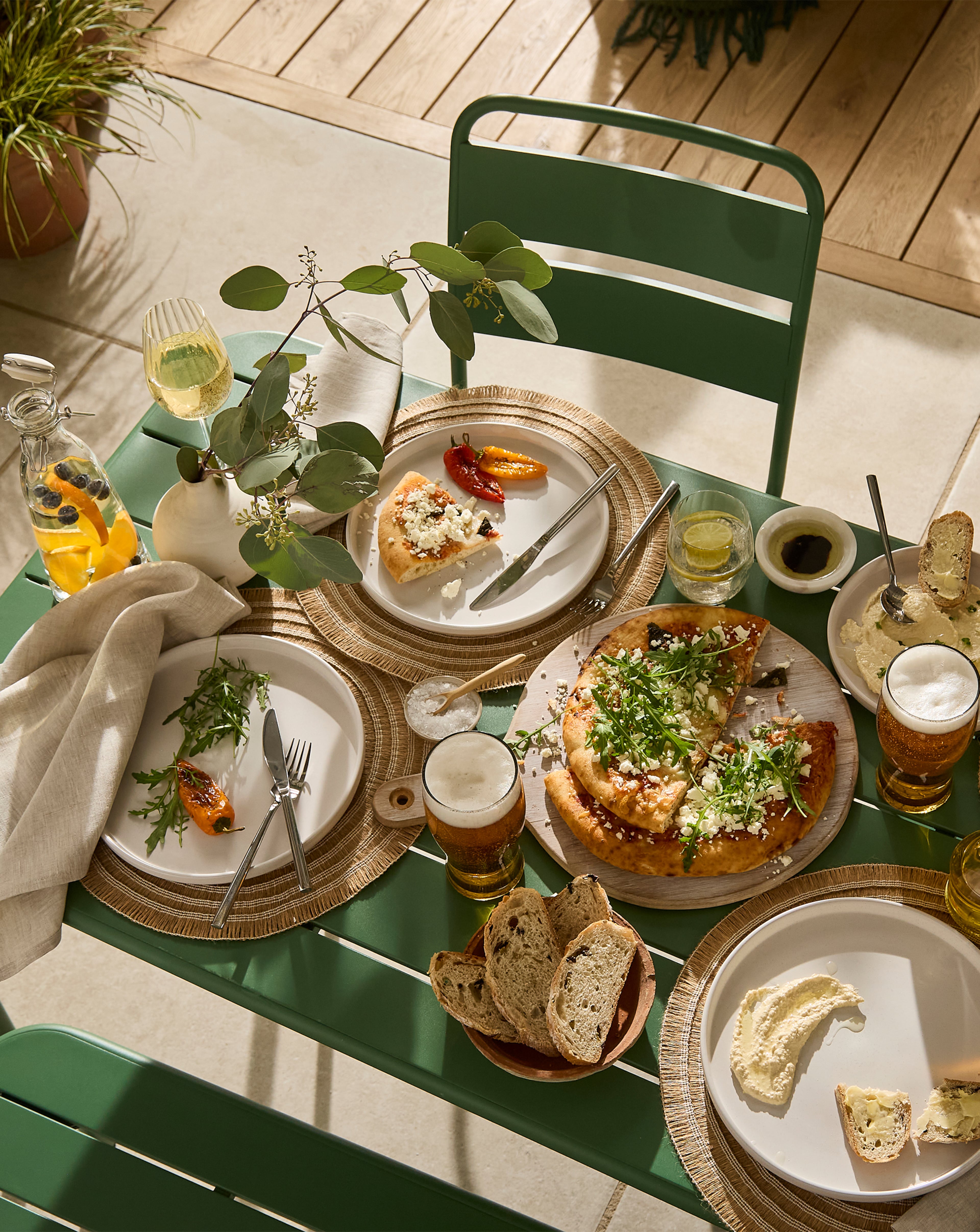 Close-up of a sunlit outdoor table set with pizza, bread, dips and drinks.