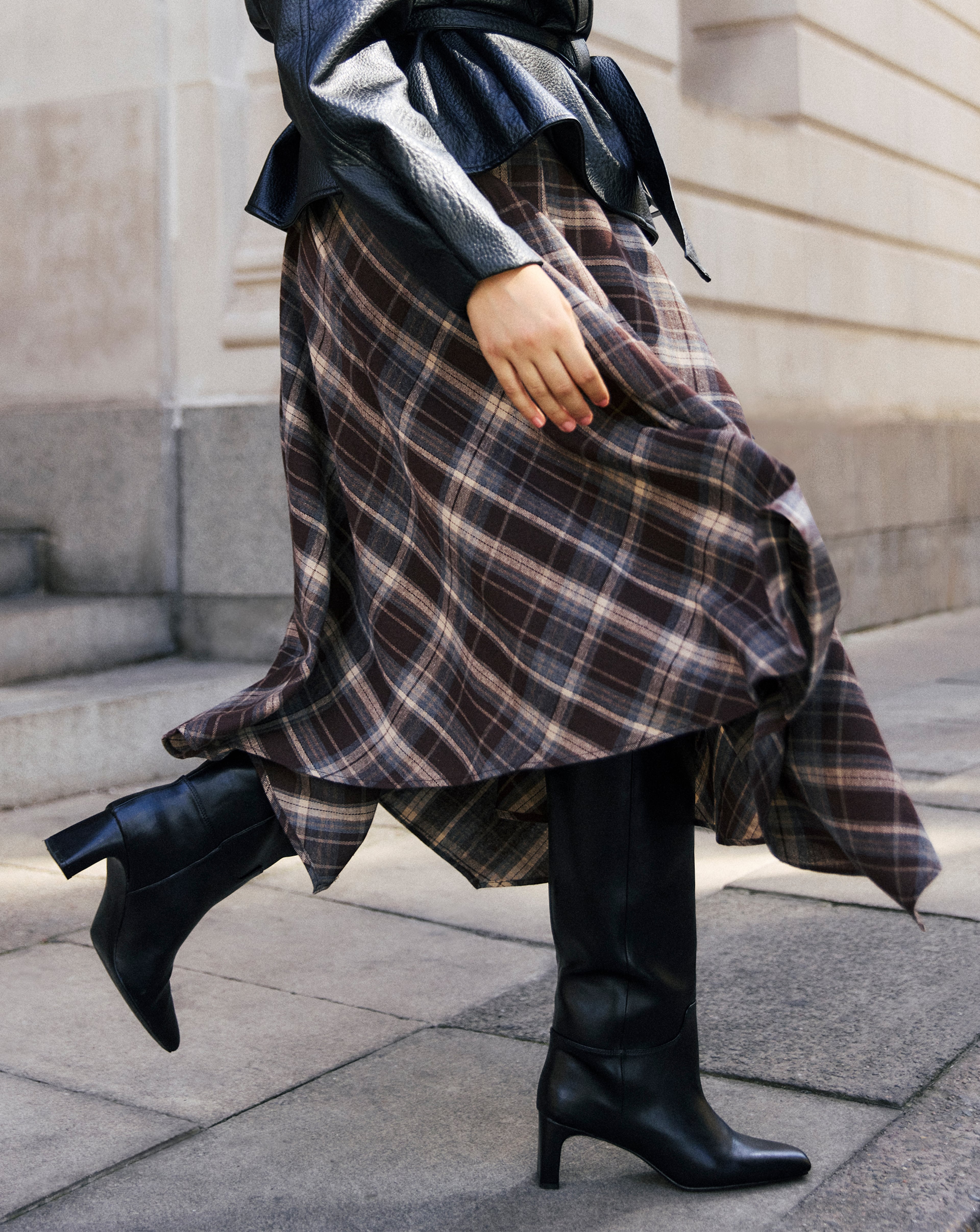 A person wearing a black leather jacket, a flowing plaid skirt, and black high-heeled boots.