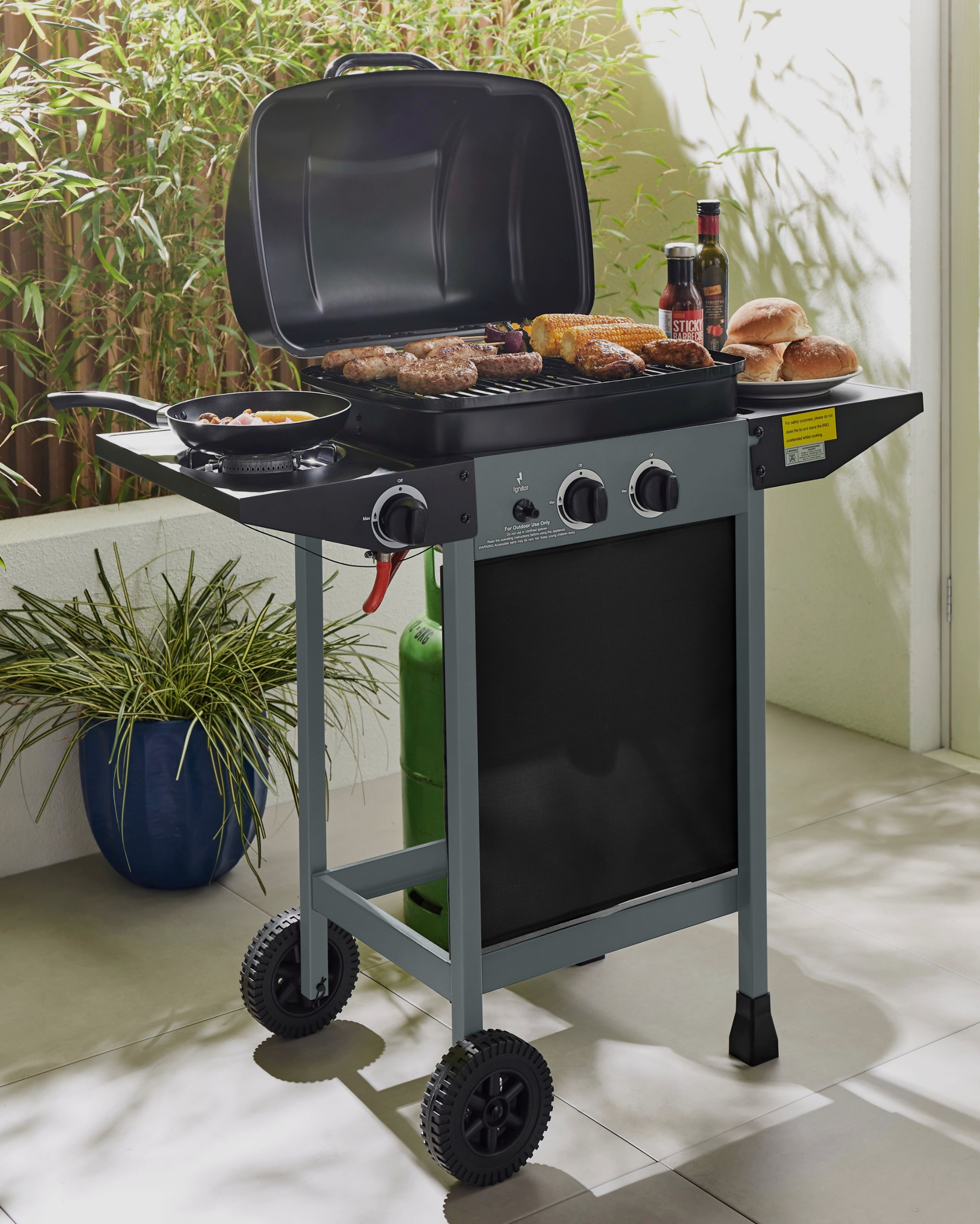 Outdoor gas barbecue grill on a patio, cooking burgers, sausages and corn, with condiments and bread rolls on the side shelves.