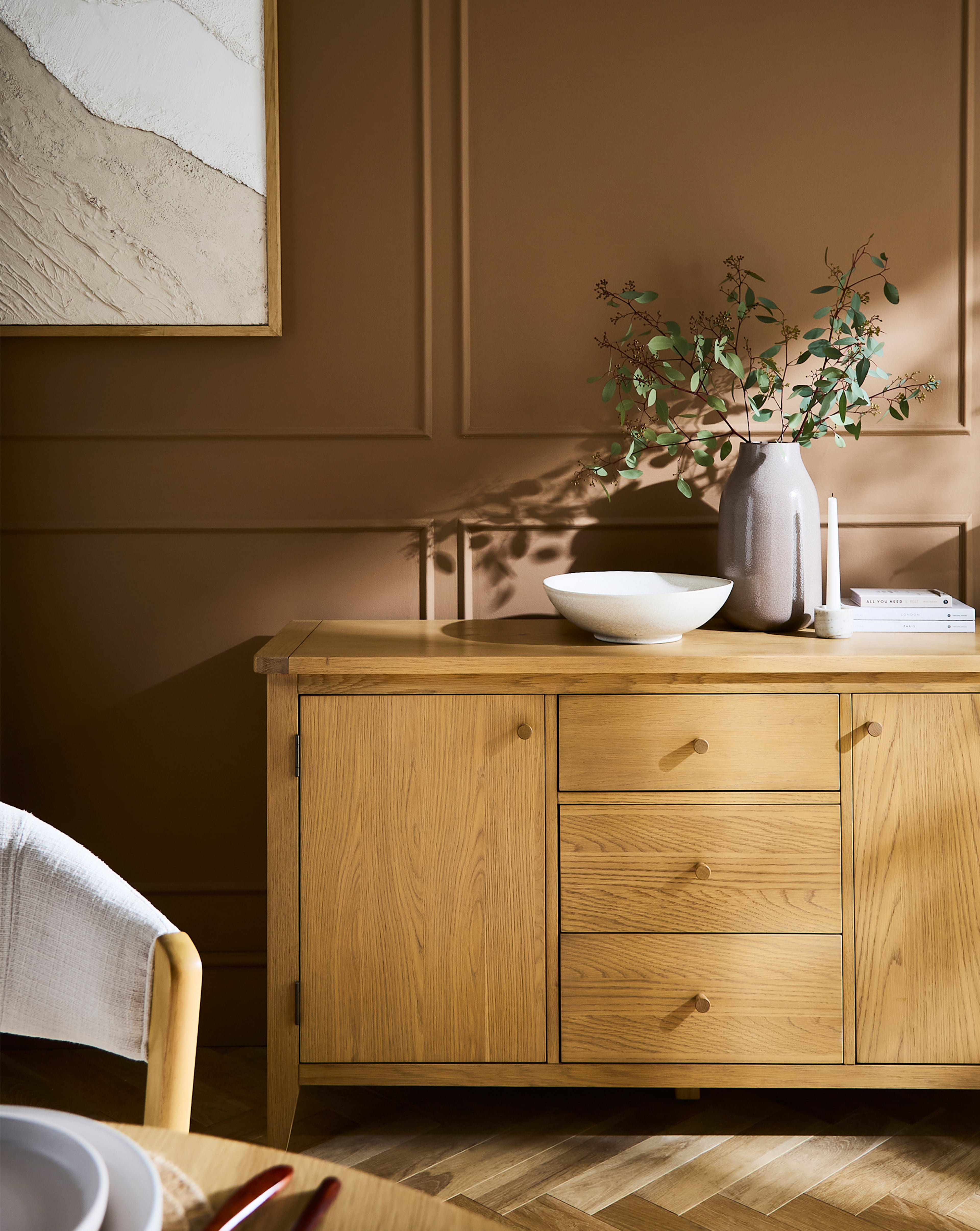 Light oak sideboard, decorated with a ceramic vase, a white bowl, and stacked books, with an abstract artwork hanging above.