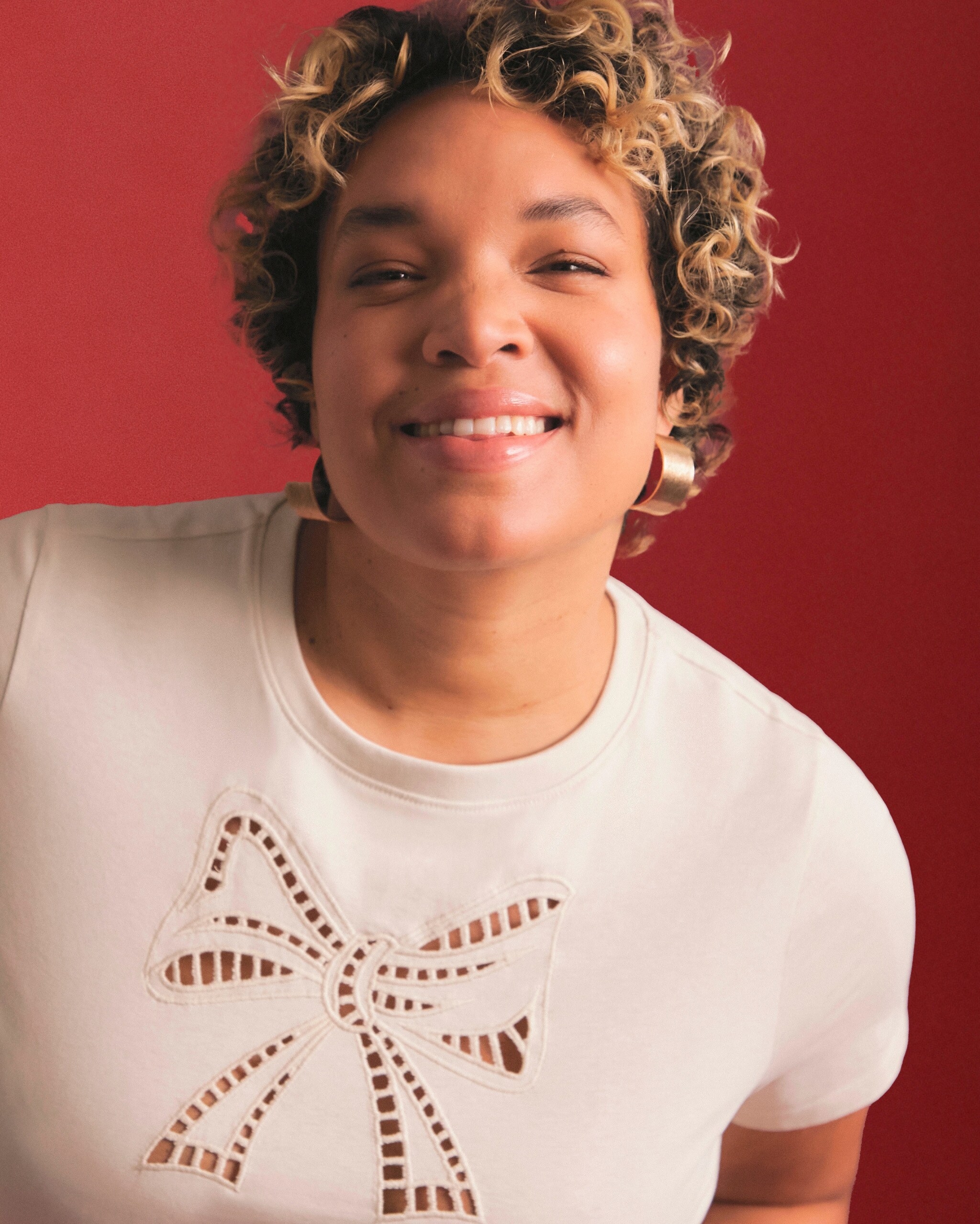 A woman with curly hair wearing a white t-shirt adorned with a bow, smiling confidently at the camera.