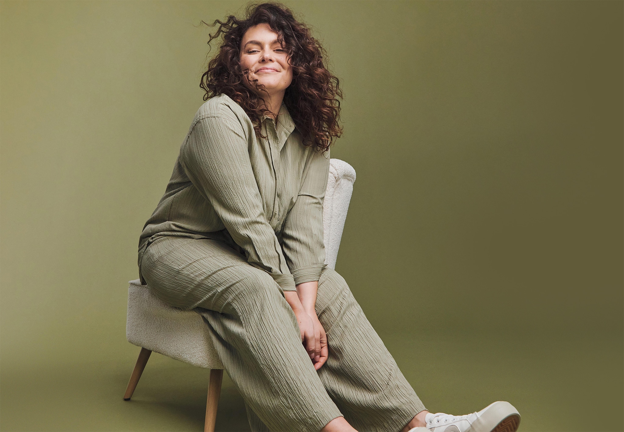 Model sat on a chair wearing an olive green loose fitting jumpsuit