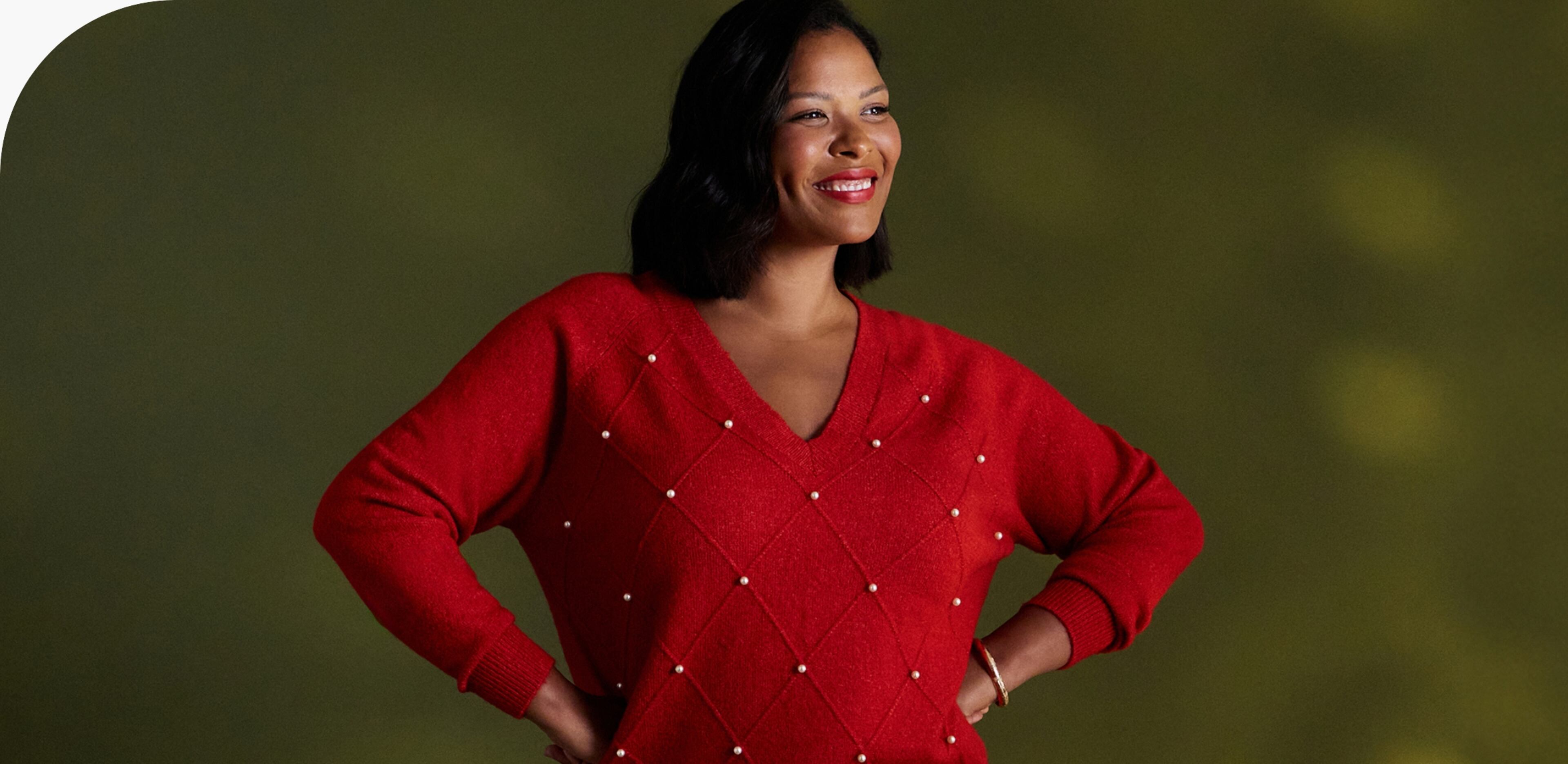 A person wearing a bright red V-neck sweater with a diamond pattern, standing against a green background with hands on hips.