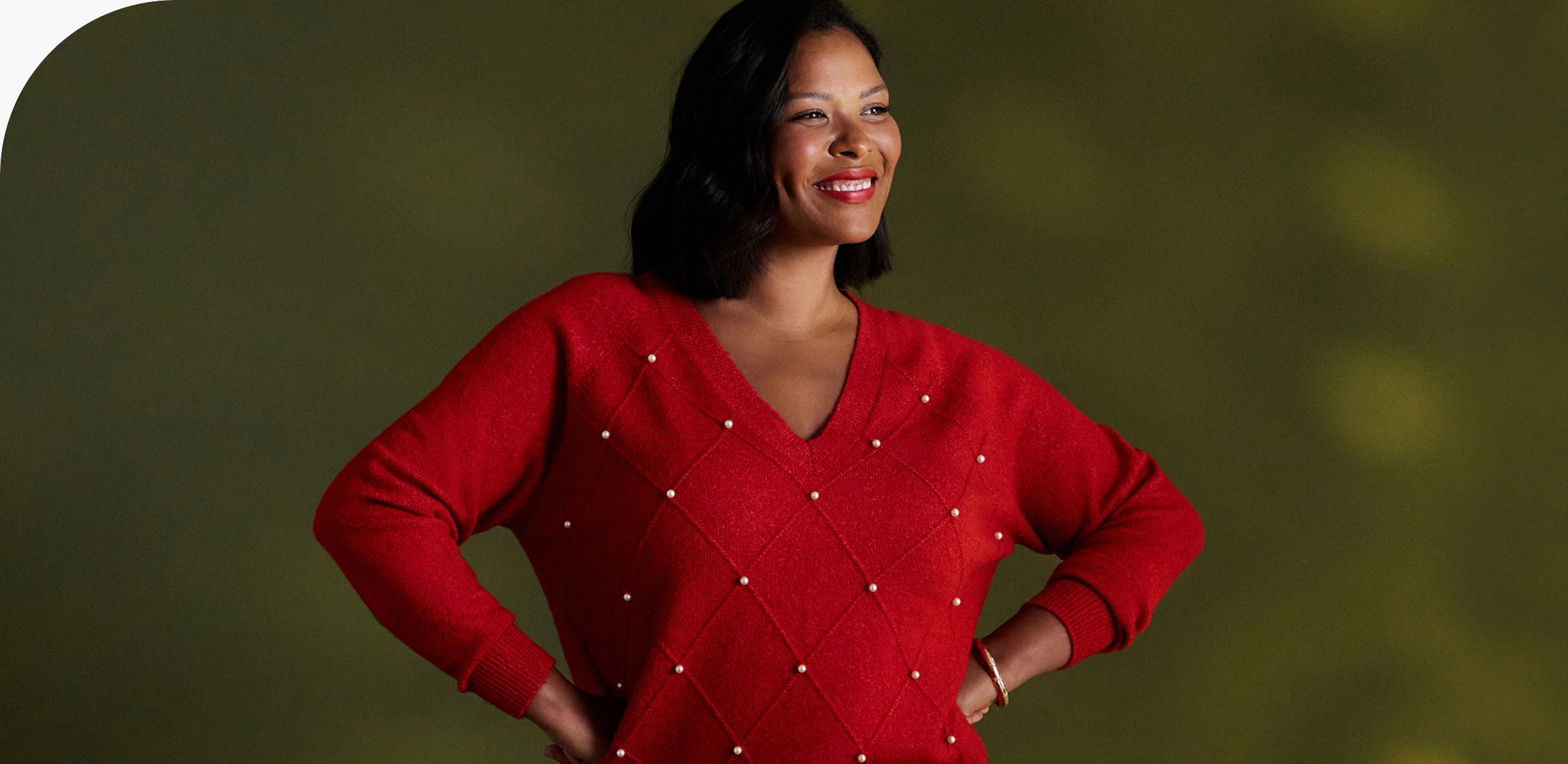 A person wearing a bright red V-neck sweater with a diamond pattern, standing against a green background with hands on hips.