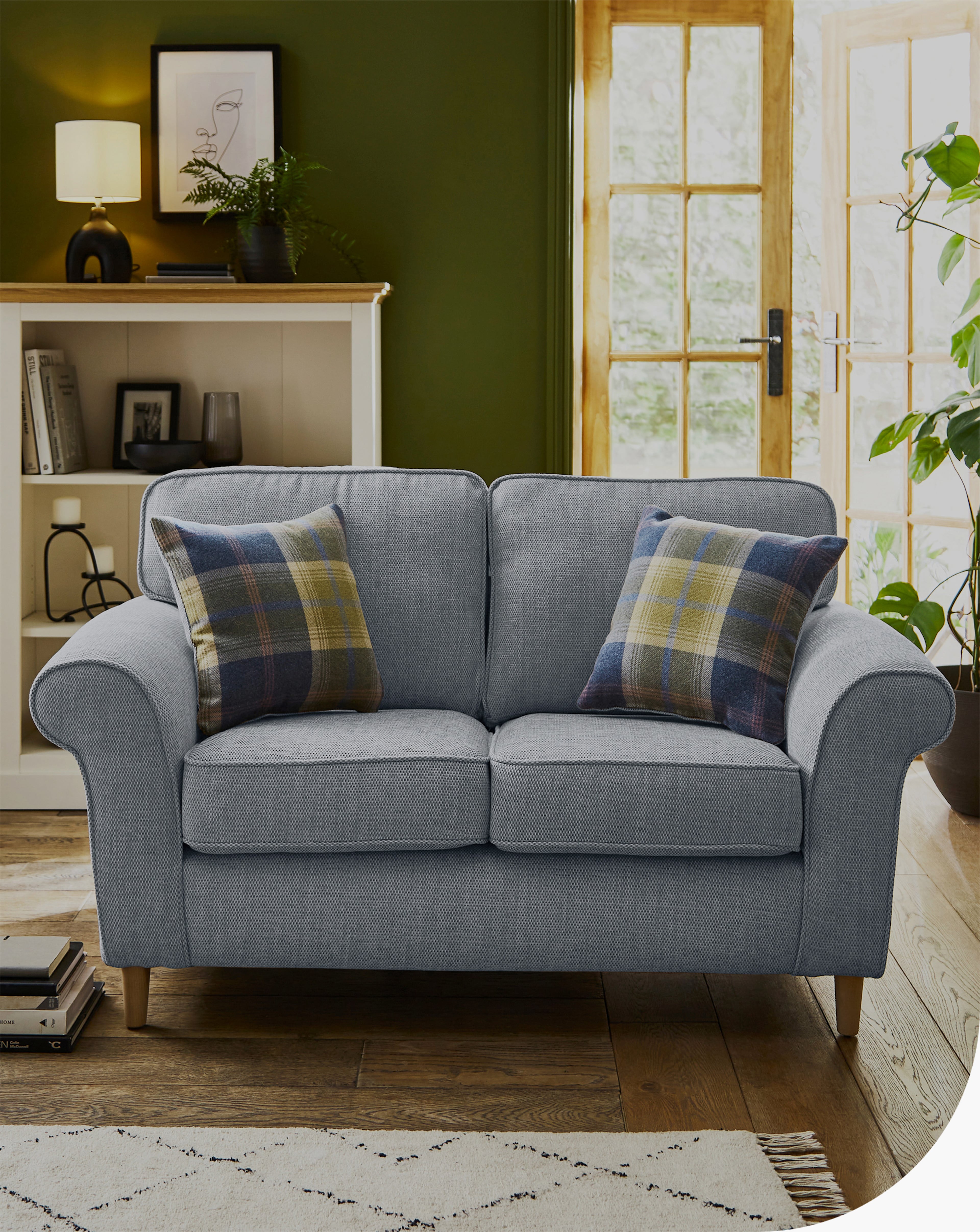 A blue-coloured Julipa Chiltern Fabric 2 Seater sofa sits in a cosy living room, decorated with two plaid cushions. Behind it is a white shelving unit with books and a lamp.