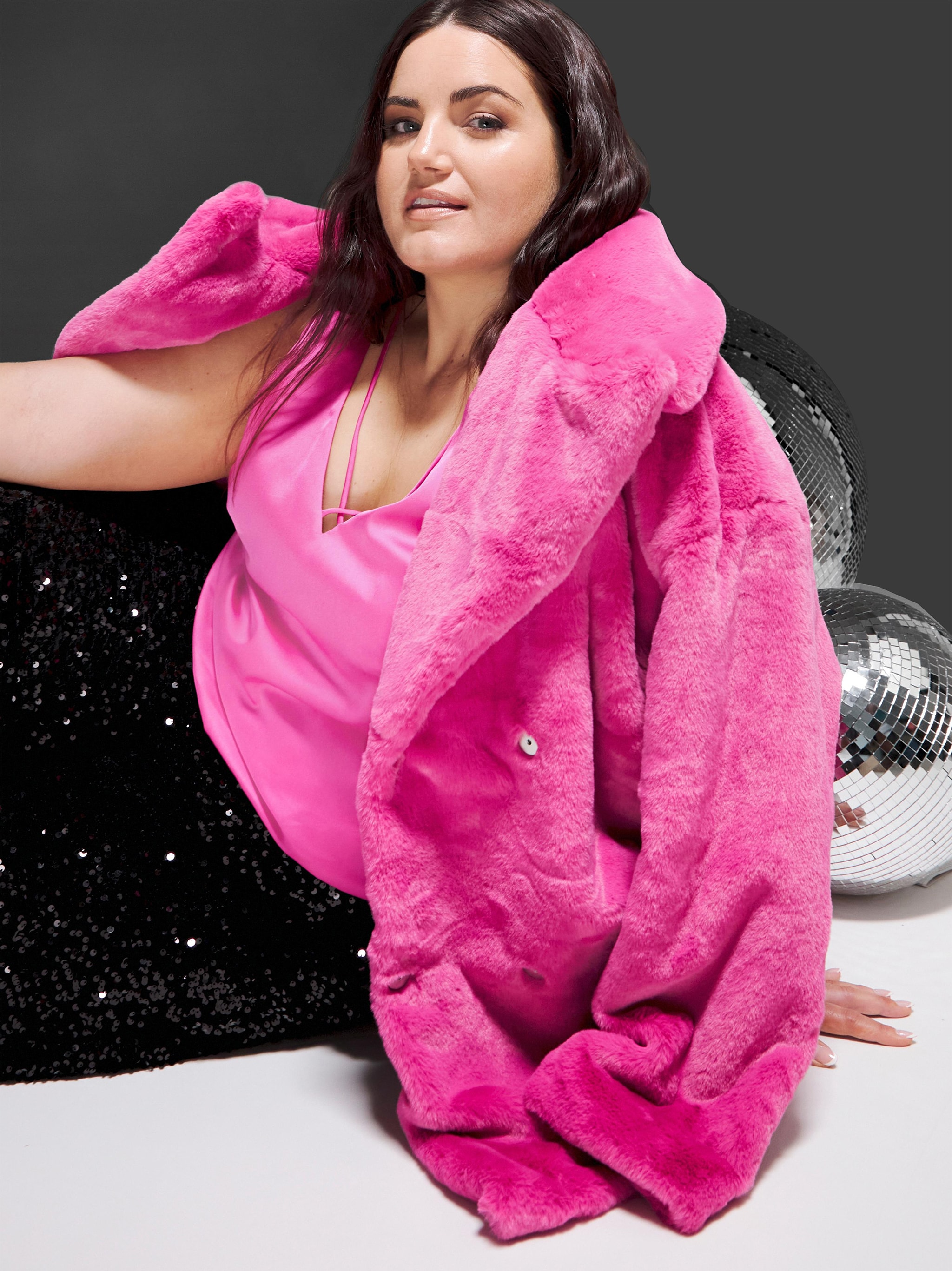 A woman sat wearing a sparkly pink vest top, pink faux fur coat, sparkly black trousers